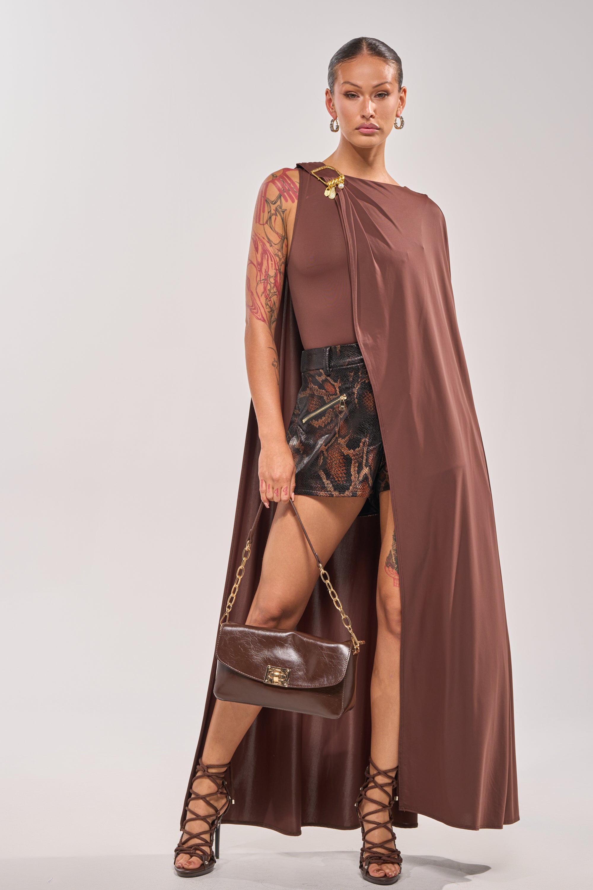 A woman stands confidently against a plain background in the NOT YOURS CAPE BODYSUIT, styled with patterned shorts, lace-up high heels, and a brown handbag with a gold buckle—a chic look for date night. 0