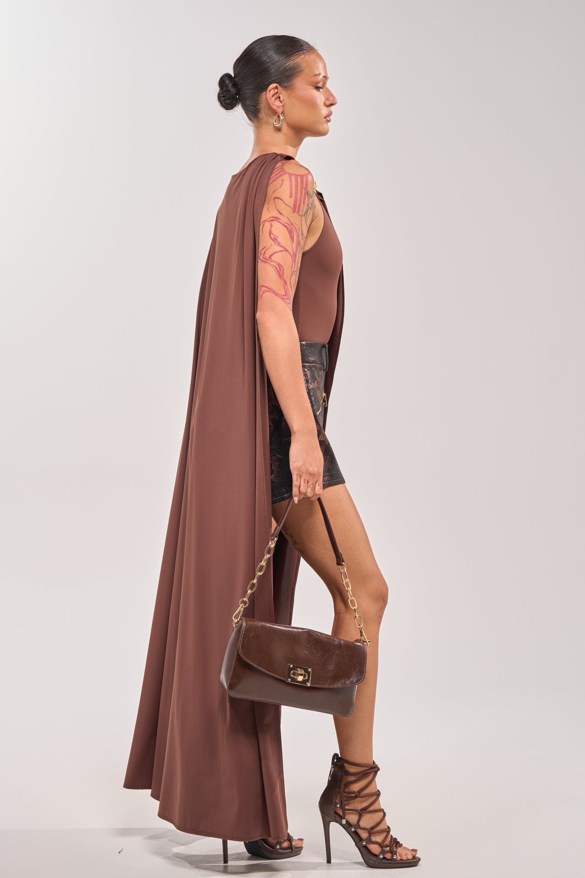 Woman in profile wears the NOT YOURS CAPE BODYSUIT in mauve, styled with black shorts, strappy heels, and a brown handbag. Her sleek bun and subtle arm tattoos pop against a plain light gray background. 3