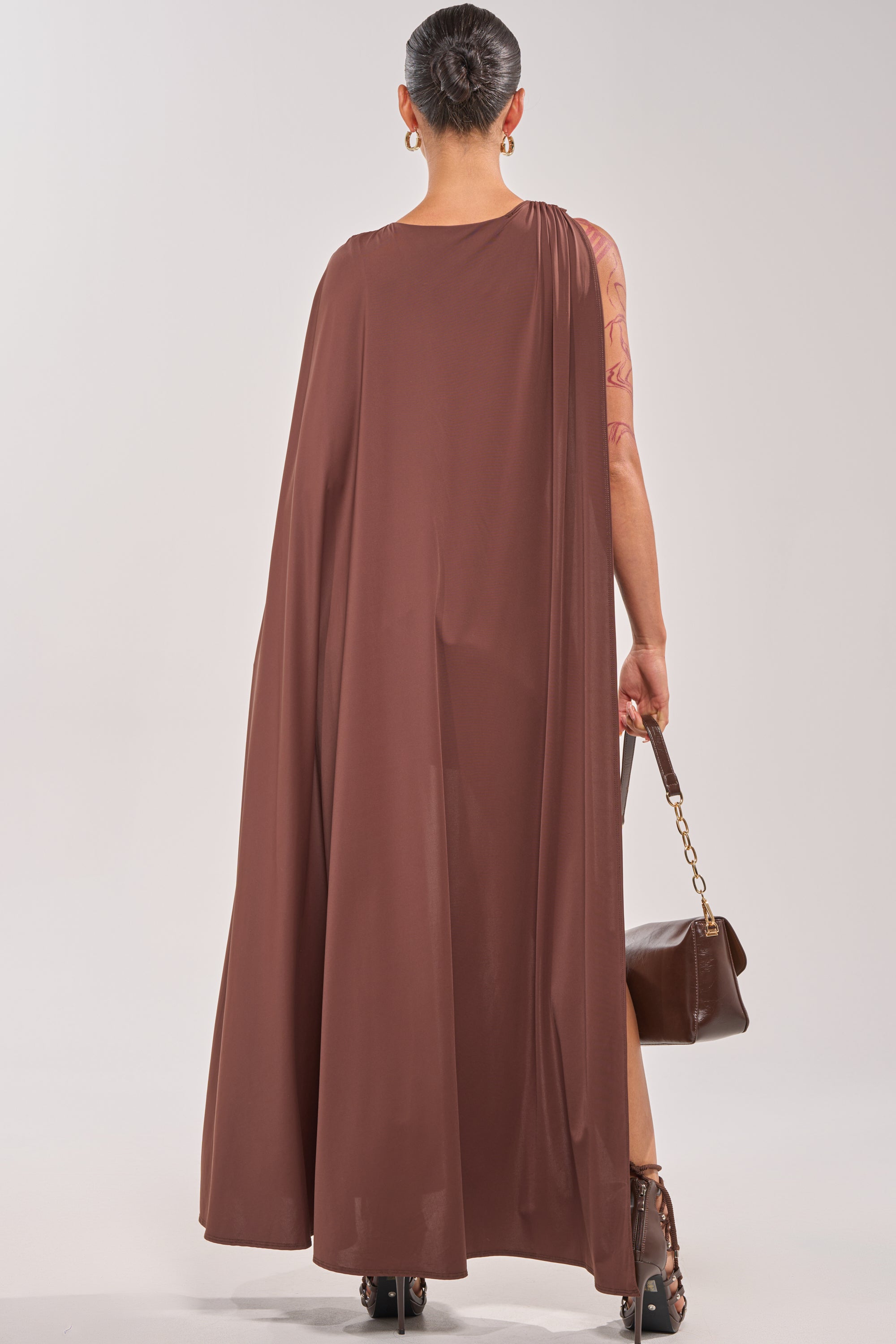 Seen from behind, a person with their hair in a bun wears the NOT YOURS CAPE BODYSUIT in brown, paired with high heels and a matching handbag. A tattooed arm is visible against a plain light background. 5