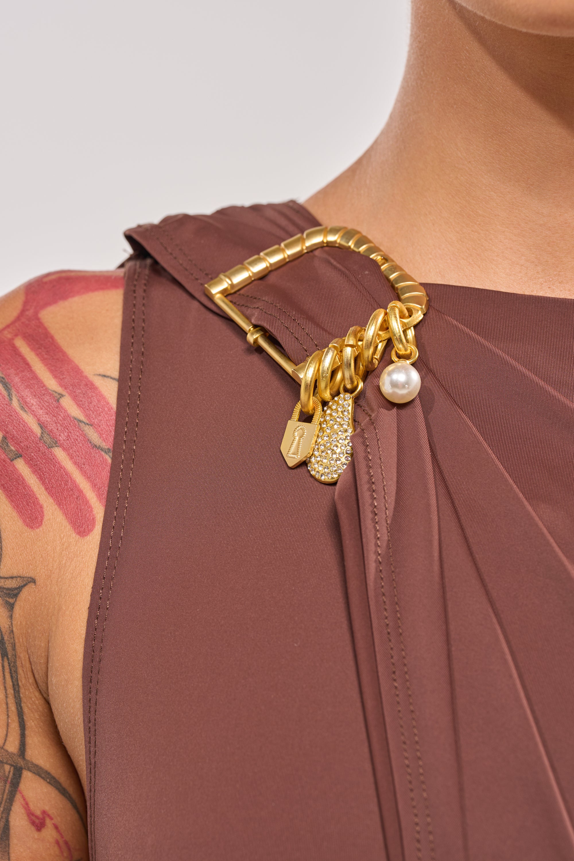 Close-up of a person in the NOT YOURS CAPE BODYSUIT, a sleeveless brown design with editorial styling. Features include a gold buckle accent and pearl charms on the shoulder. A red tattoo is partly visible on the upper arm. 6
