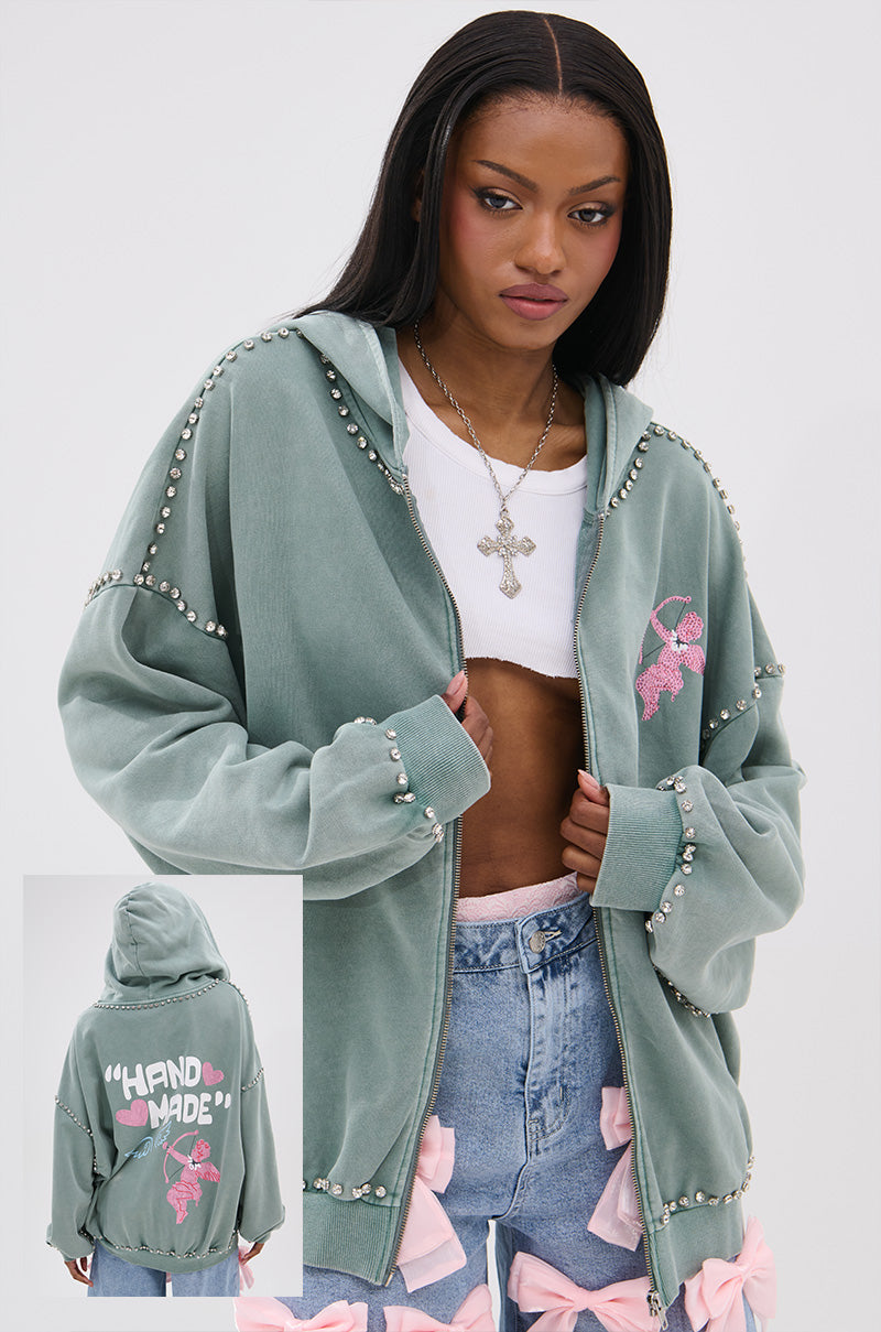 A woman wears the HAND MADE ZIP UP SWEATSHIRT, an oversized sage green hoodie with pearl, pink bows, rhinestone graphics, and “HAND MADE” on the back. She pairs it with a white crop top, jeans with pink bows, and straight black hair.