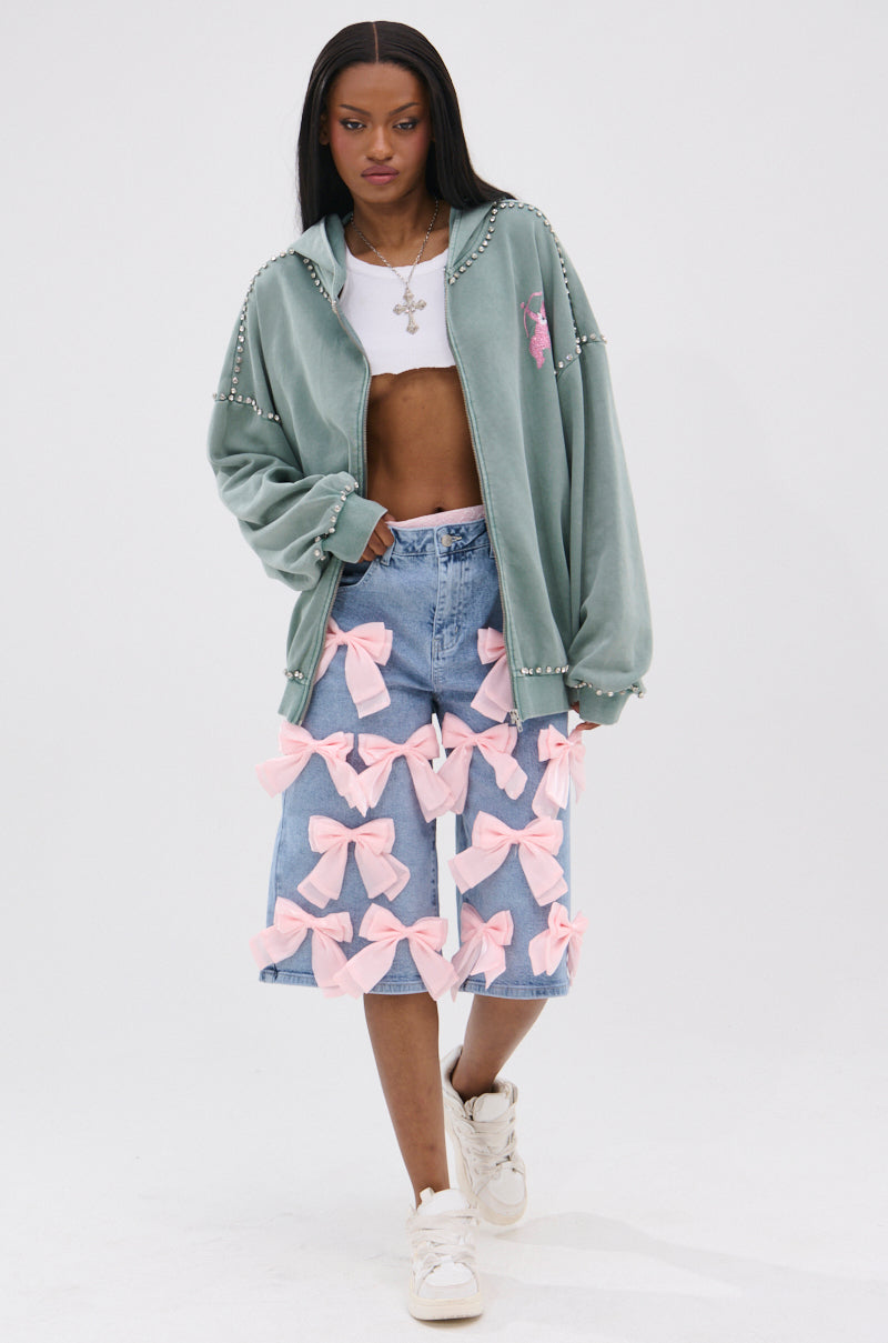 A woman wears the HAND MADE ZIP UP SWEATSHIRT in sage green, paired with a white crop top and a denim skirt adorned with large pink bows. She finishes her look with white sneakers, a silver necklace, standing against a plain white background.