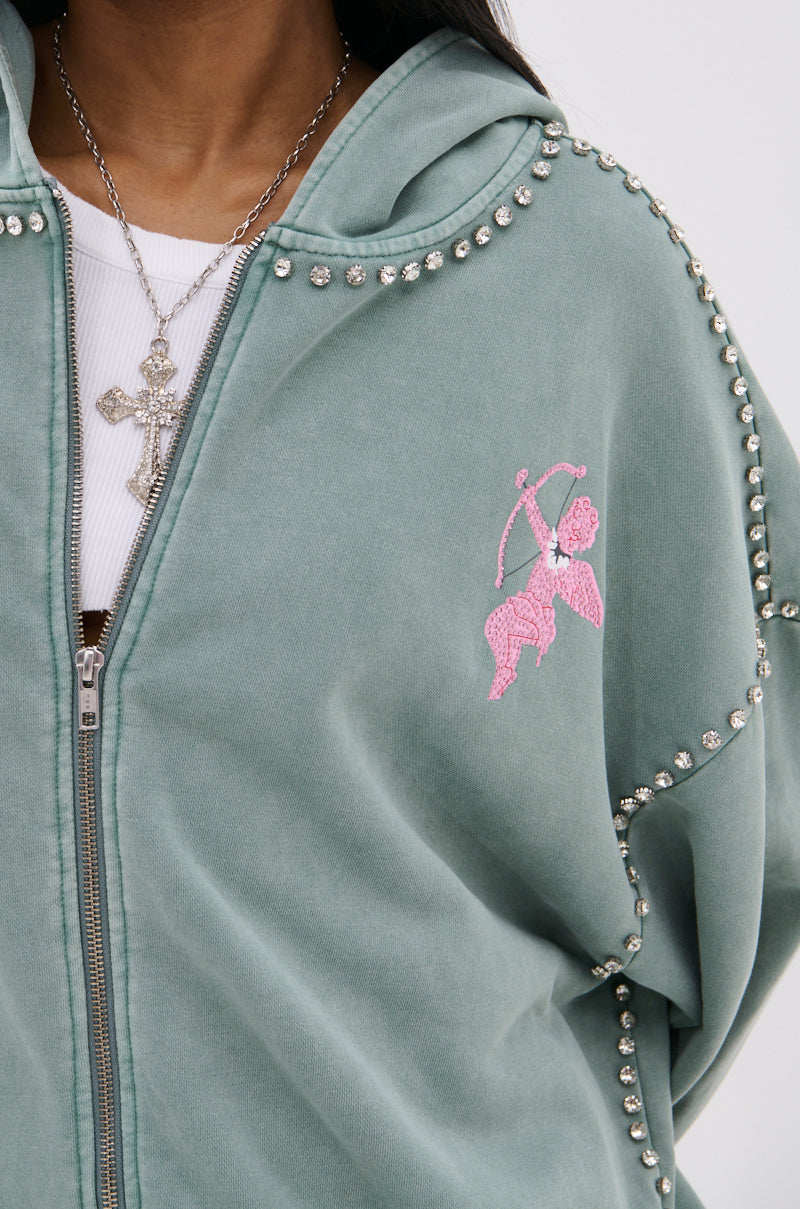 A close-up of a person wearing the HAND MADE ZIP UP SWEATSHIRT—an oversized green zip-up hoodie with silver studs on the seams and a pink embroidered Sagittarius zodiac sign—styled with a silver cross necklace layered over a white top.