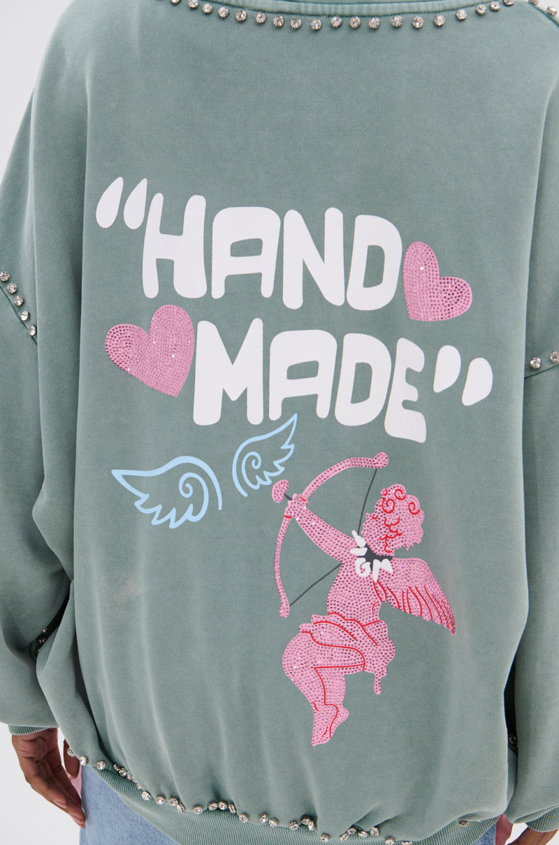 Wearing a HAND MADE ZIP UP SWEATSHIRT, featuring green fabric, bold white “HAND MADE” text, pink hearts, blue wings, and a rhinestone-studded pink cherub with bow and arrow graphic on the back for standout style.