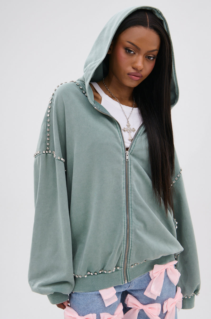 A woman with long straight hair wears the HAND MADE ZIP UP SWEATSHIRT, an AKIRA Label sage green oversized hoodie with pearl embellishments, a white top, and light blue jeans with large pink bows. She stands against a plain white background.