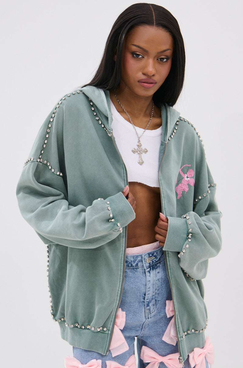A woman wears an oversized HAND MADE ZIP UP SWEATSHIRT in light green with pearl-like studs, a white crop top, high-waisted blue jeans with pink bows, straight dark hair, and a large cross necklace.