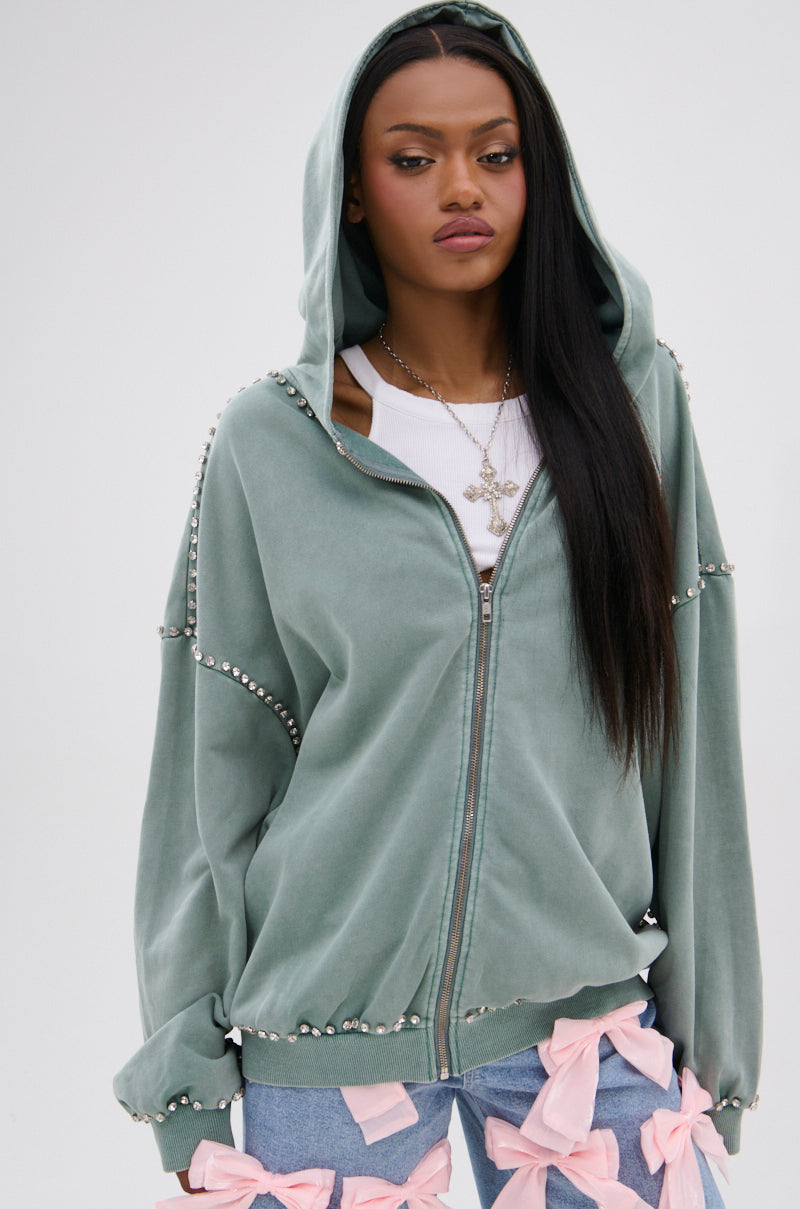 A woman wears the HAND MADE ZIP UP SWEATSHIRT, an oversized sage green zip-up hoodie with rhinestone graphics, over a white top and blue jeans with pink bows, accessorized with a cross necklace against a plain background.