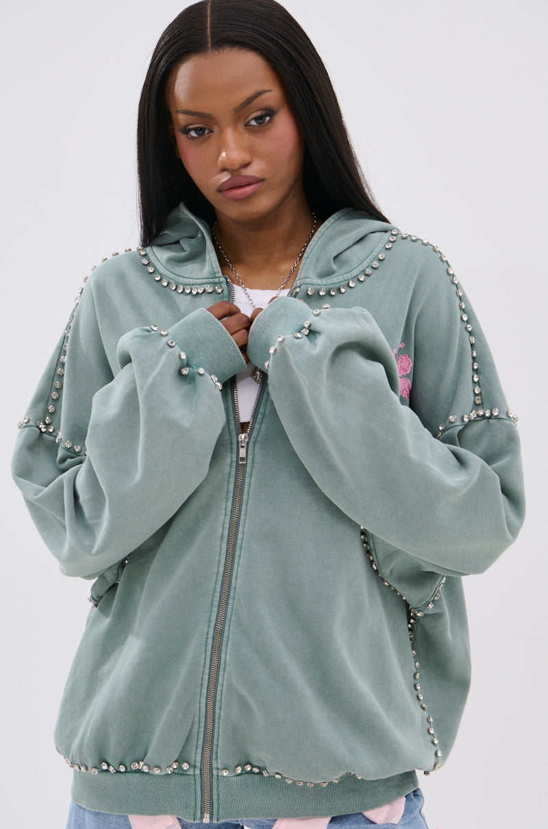 A woman with long straight hair wears an oversized, sage green HAND MADE ZIP UP SWEATSHIRT with rhinestone graphics over a white t-shirt, standing against a plain light background.
