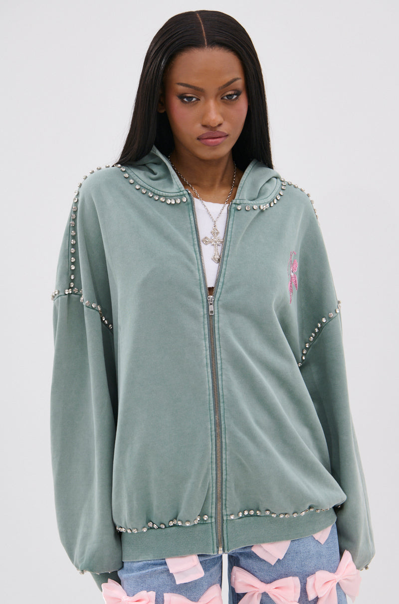 A woman wears the HAND MADE ZIP UP SWEATSHIRT in sage green with pearl embellishments and a small pink chest detail, paired with denim shorts featuring pink bows, creating a chic and playful look against a plain background.