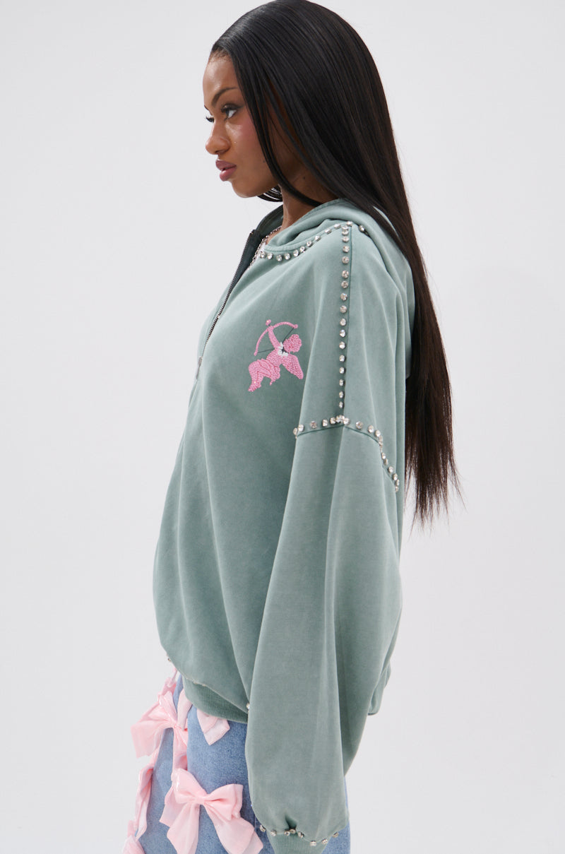 A woman with long straight hair wears the AKIRA Label Hand Made Zip Up Sweatshirt—an oversized, sage green velour hoodie with silver studs, rhinestone cupid graphics on the sleeve—and blue jeans with large pink bows.