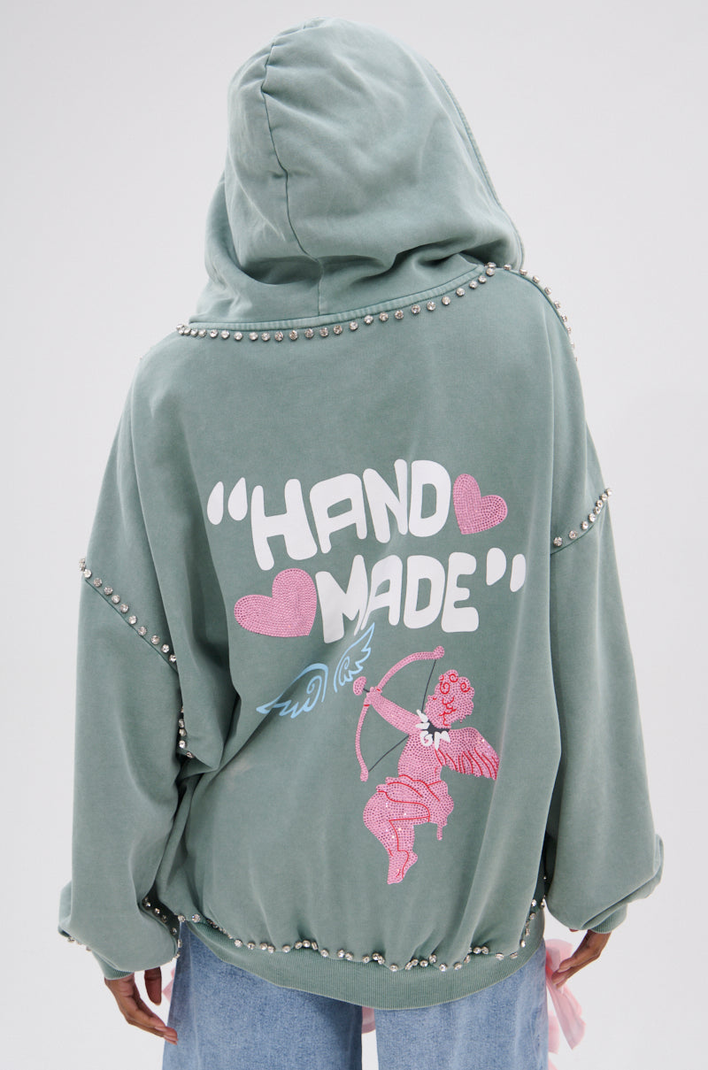 A person wears the oversized HAND MADE ZIP UP SWEATSHIRT from AKIRA Label in sage green, featuring rhinestone graphics and a back design with "HAND MADE" text and a pink cherub aiming a bow, surrounded by pink hearts.
