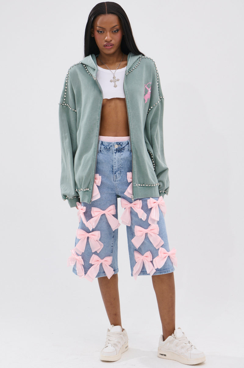 A woman stands against a white background wearing the HAND MADE ZIP UP SWEATSHIRT in sage green with rhinestone graphics, a white crop top, long denim shorts featuring large pink bows, white sneakers, and a silver necklace.