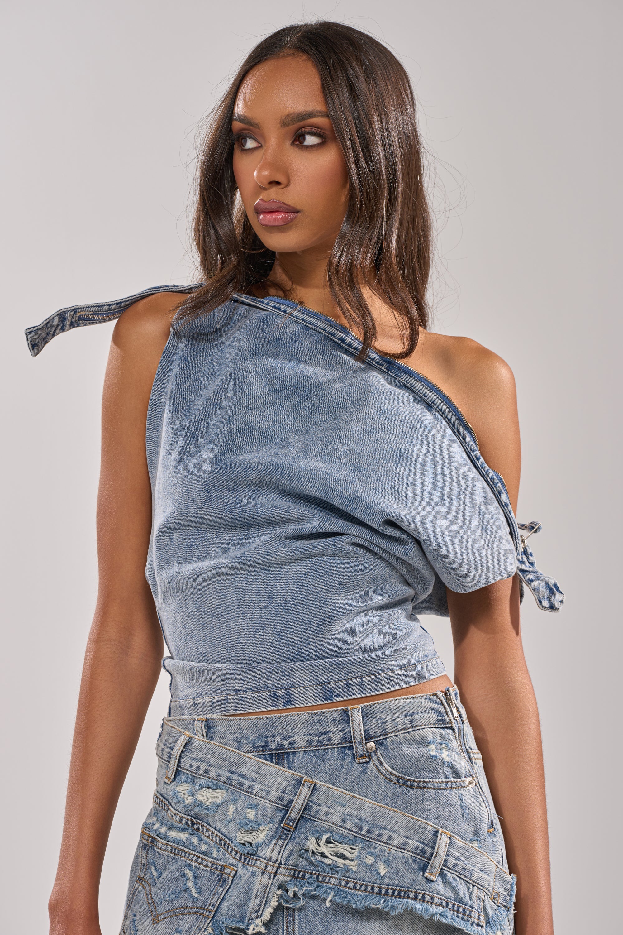 A woman wearing the WEEKEND VIBES DENIM BLOUSE and a frayed denim skirt stands against a neutral background, gazing to her left. 0