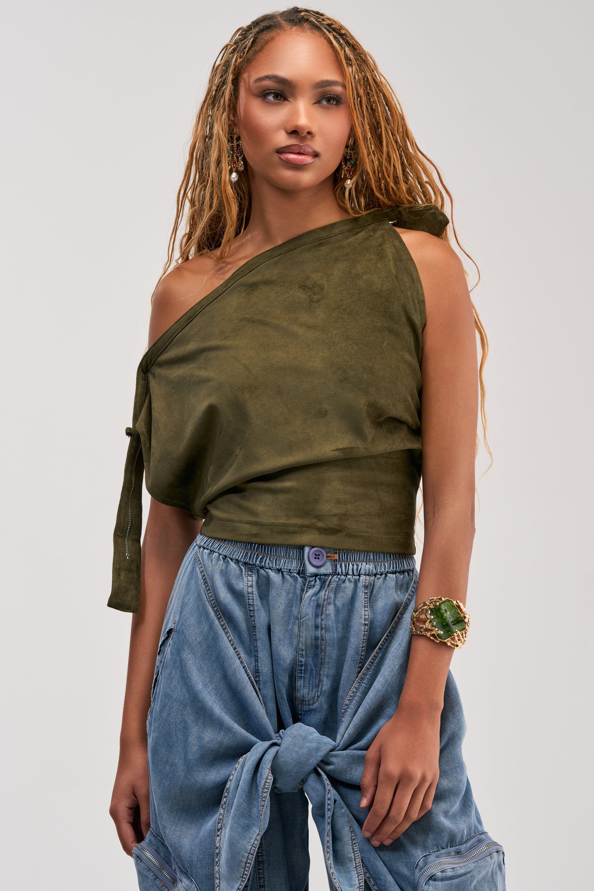 A woman stands against a plain background wearing the GOOD VIBES ONLY SUEDE OFF SHOULDER TOP in olive green, paired with loose blue jeans featuring a large front tie, and accessorized with a green bracelet and earrings.