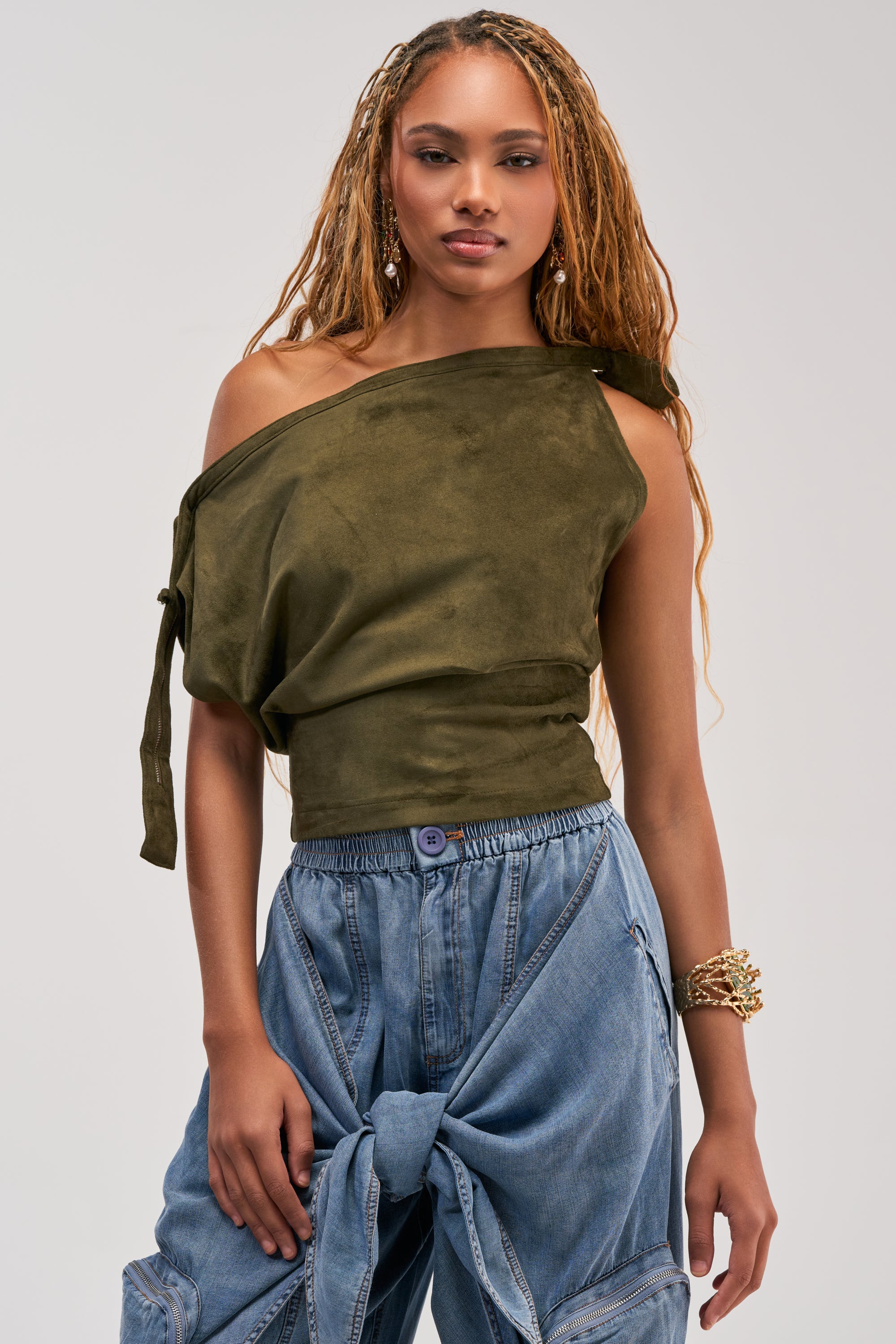 Wearing the GOOD VIBES ONLY SUEDE OFF SHOULDER TOP in olive, a woman with long braids and loose blue jeans knotted at the waist poses confidently against a plain background; the blouse features subtle zipper details for added style.