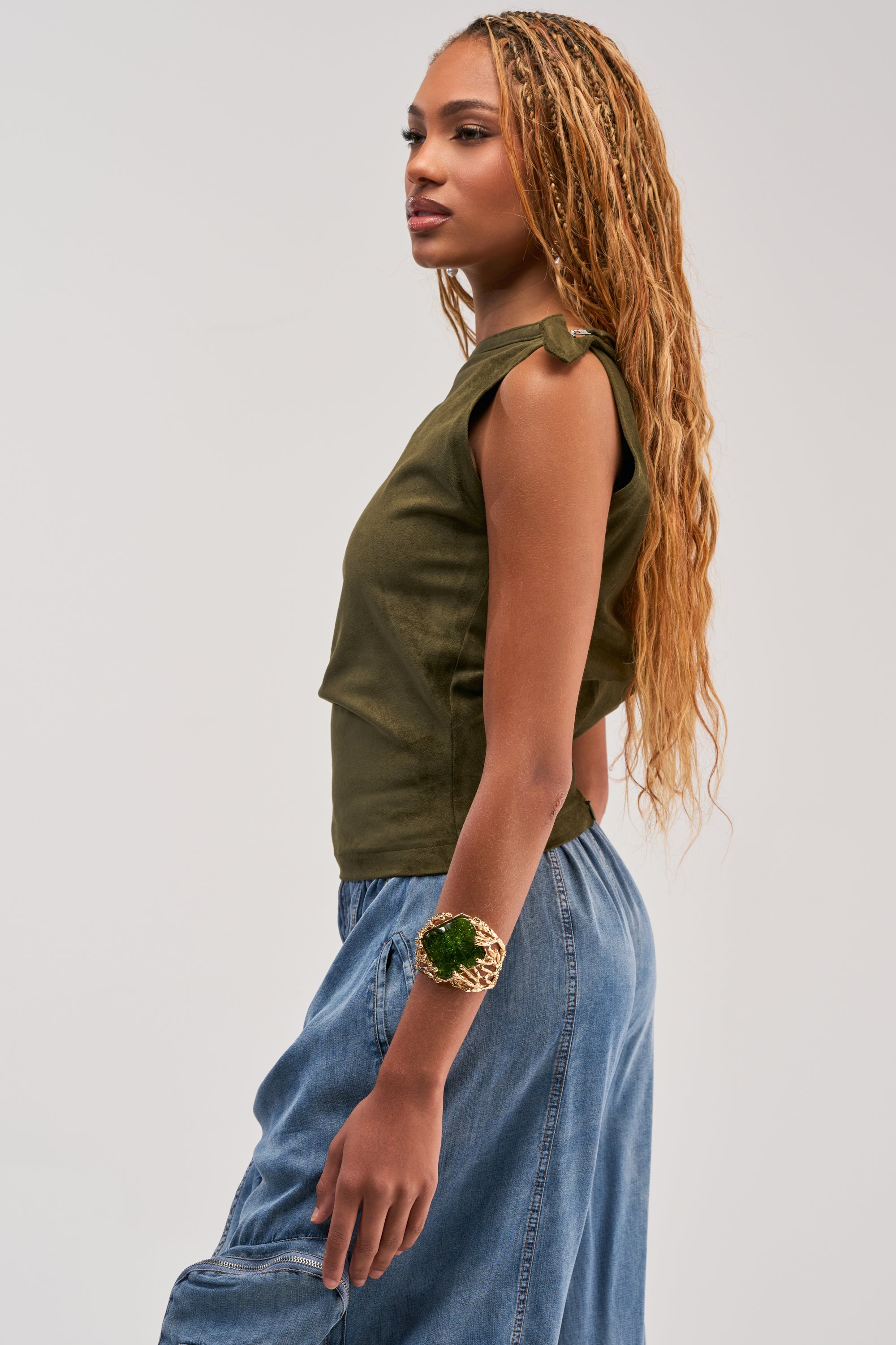 A woman with long braided hair wears the GOOD VIBES ONLY SUEDE OFF SHOULDER TOP in olive, featuring a zipper detail, paired with a chunky bracelet and blue jeans, standing sideways against a plain light background.