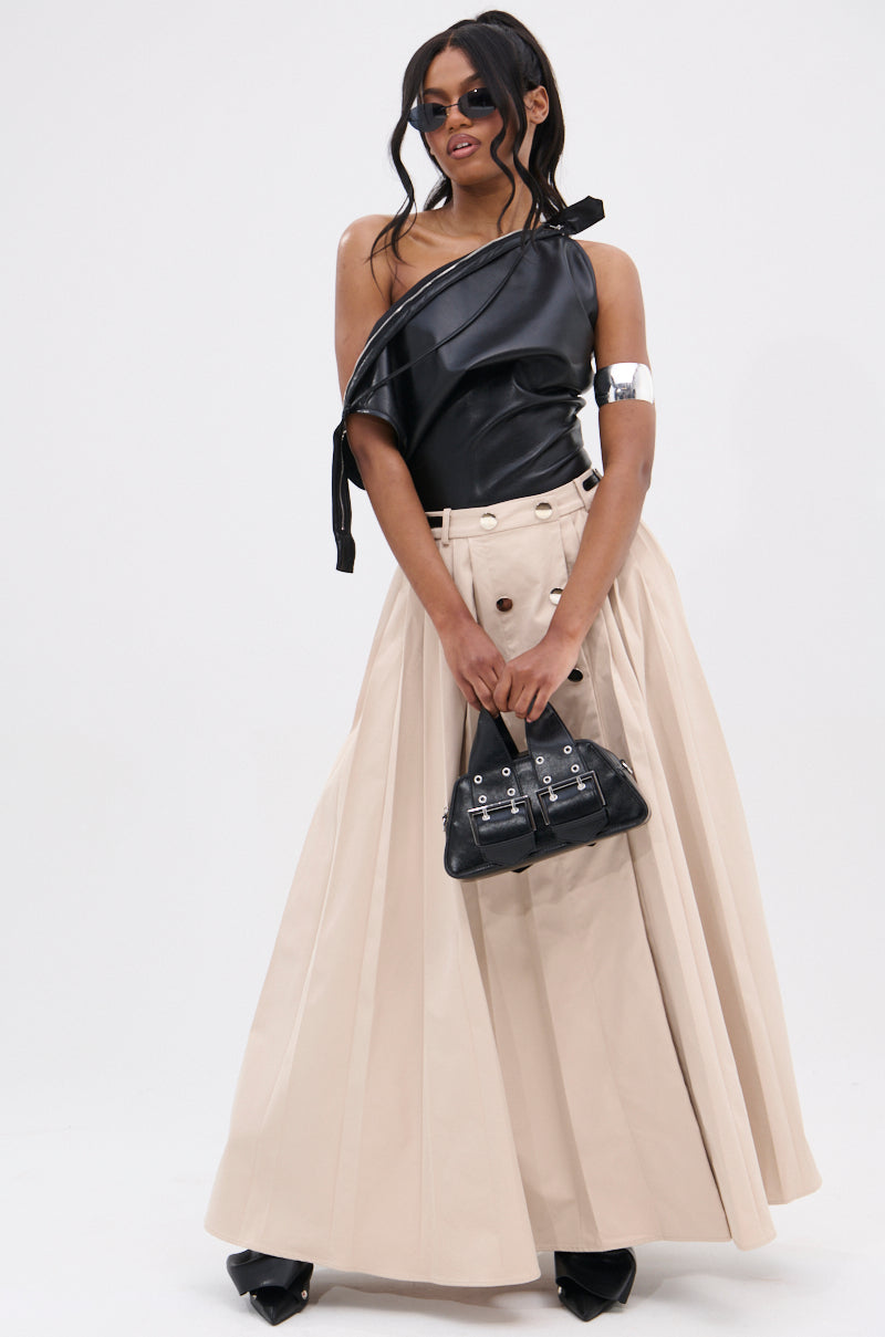 A woman in sunglasses, a black WEEKEND VIBES FAUX LEATHER BLOUSE, wide cream pleated skirt, pointed black shoes, silver arm cuff, and holding a small black handbag stands against a plain white background.