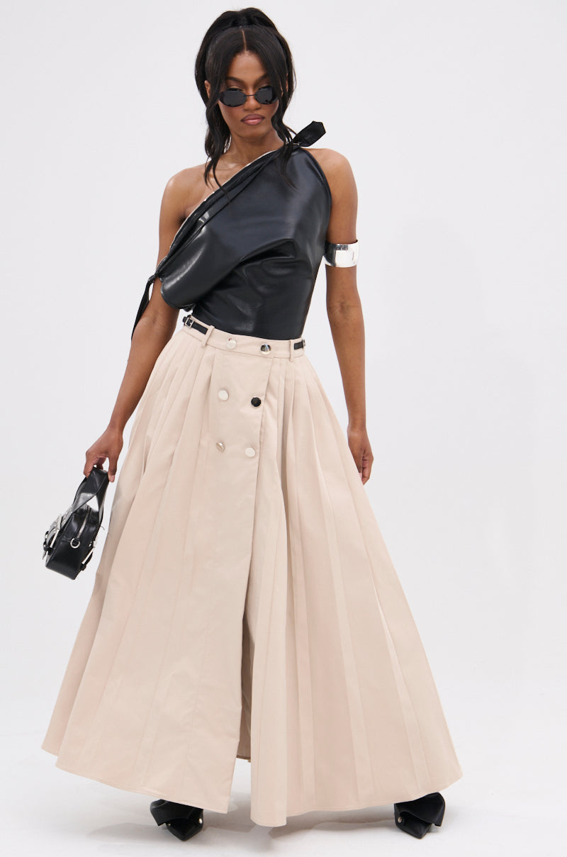 A woman wears the WEEKEND VIBES FAUX LEATHER BLOUSE, beige high-waisted pleated wide-leg pants with buttons, black pointed shoes, sunglasses, and carries a small black handbag. Her hair is styled in a low ponytail.