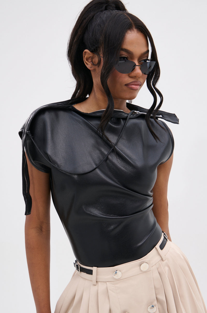 A woman in black cat-eye sunglasses, a WEEKEND VIBES FAUX LEATHER BLOUSE—an asymmetric off-the-shoulder shiny black top—and high-waisted beige pleated pants with buttons and a belt, stands against a plain light background.