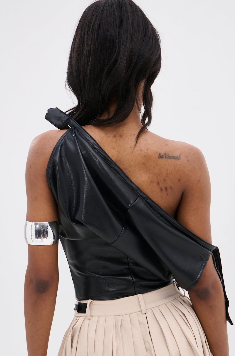 A person faces away, wearing the WEEKEND VIBES FAUX LEATHER BLOUSE in black and a beige pleated skirt. They have a visible upper arm cuff, an upper back shoulder tattoo, and long dark hair.