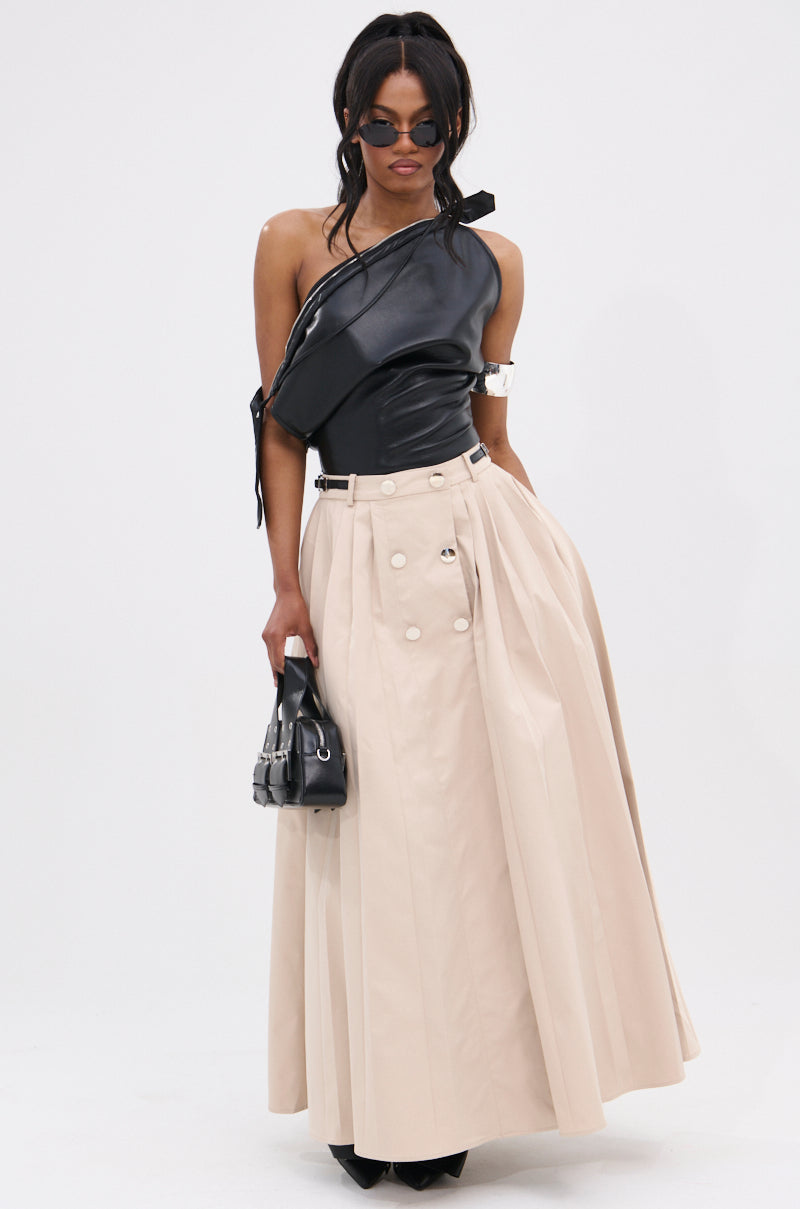 A woman in the WEEKEND VIBES FAUX LEATHER BLOUSE, featuring an asymmetric off-shoulder design, pairs it with a beige pleated skirt, black pointed shoes, sunglasses, and a black handbag against a plain white background.