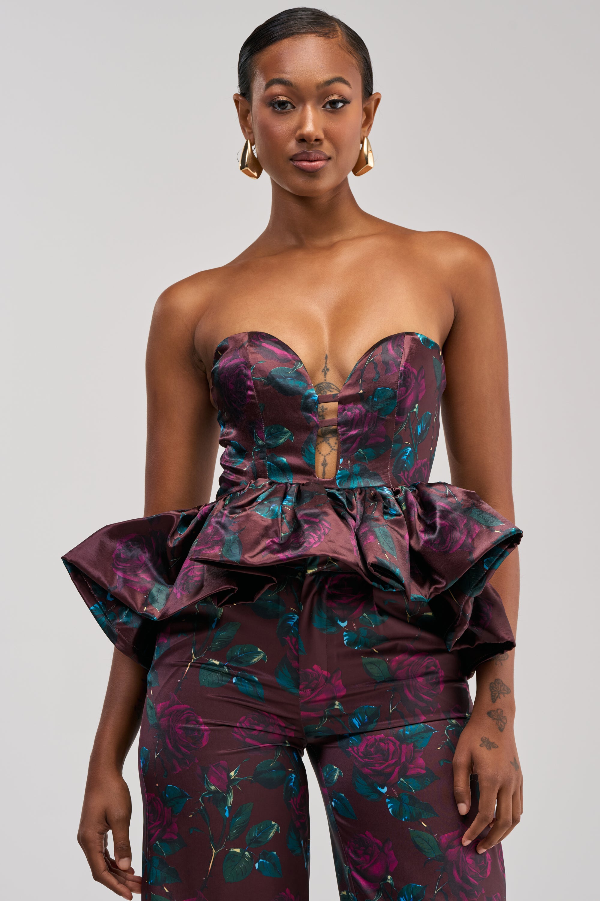 Wearing the ANTIQUE FLORAL CORSET TOP, this woman pairs the strapless, floral print peplum top with matching pants and gold hoop earrings, posing against a plain background.