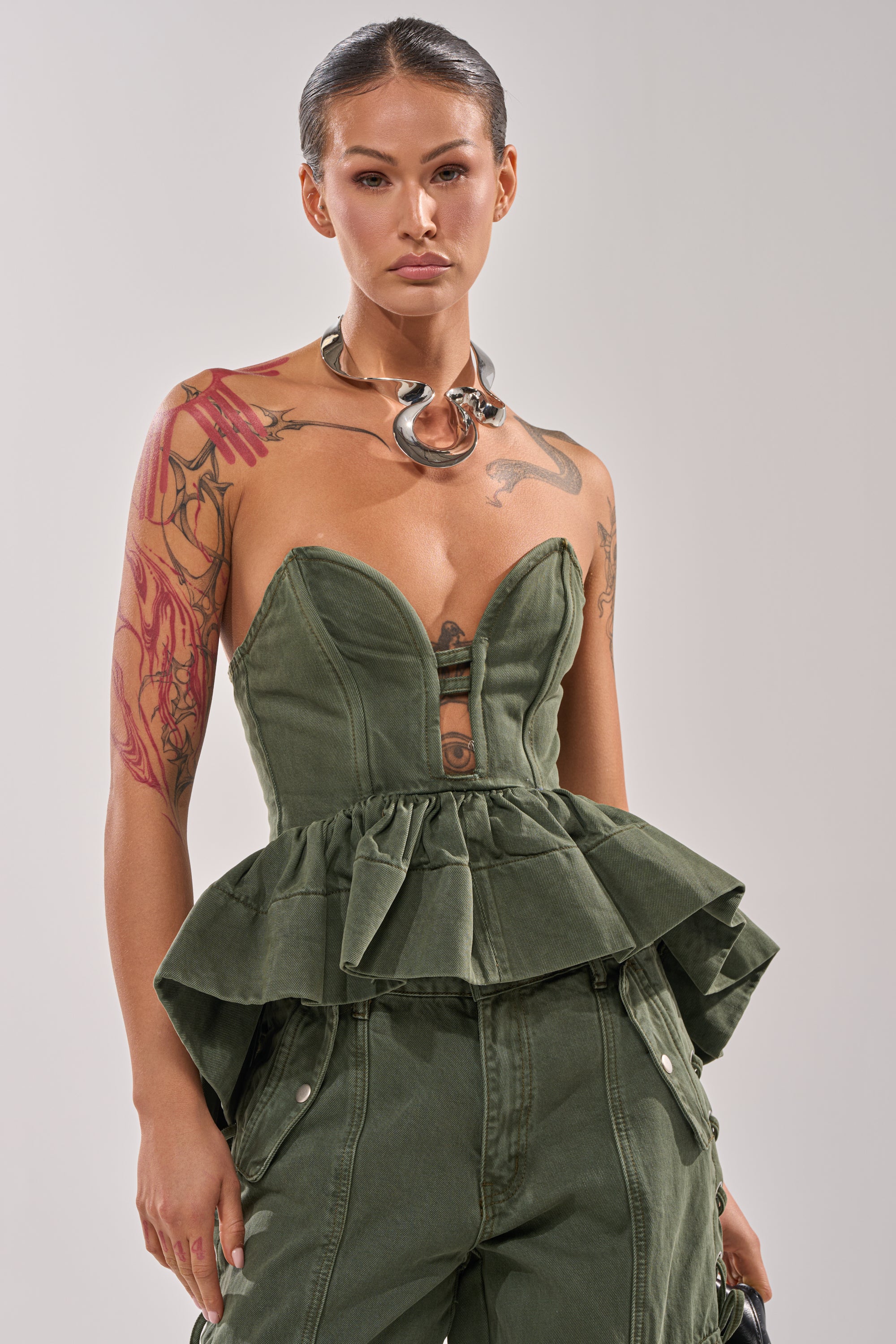 A woman with slicked-back hair models the KARA DENIM CORSET and matching pants in olive green, showing her left arm tattoos and styling a bold silver statement necklace against a plain light gray background. 0