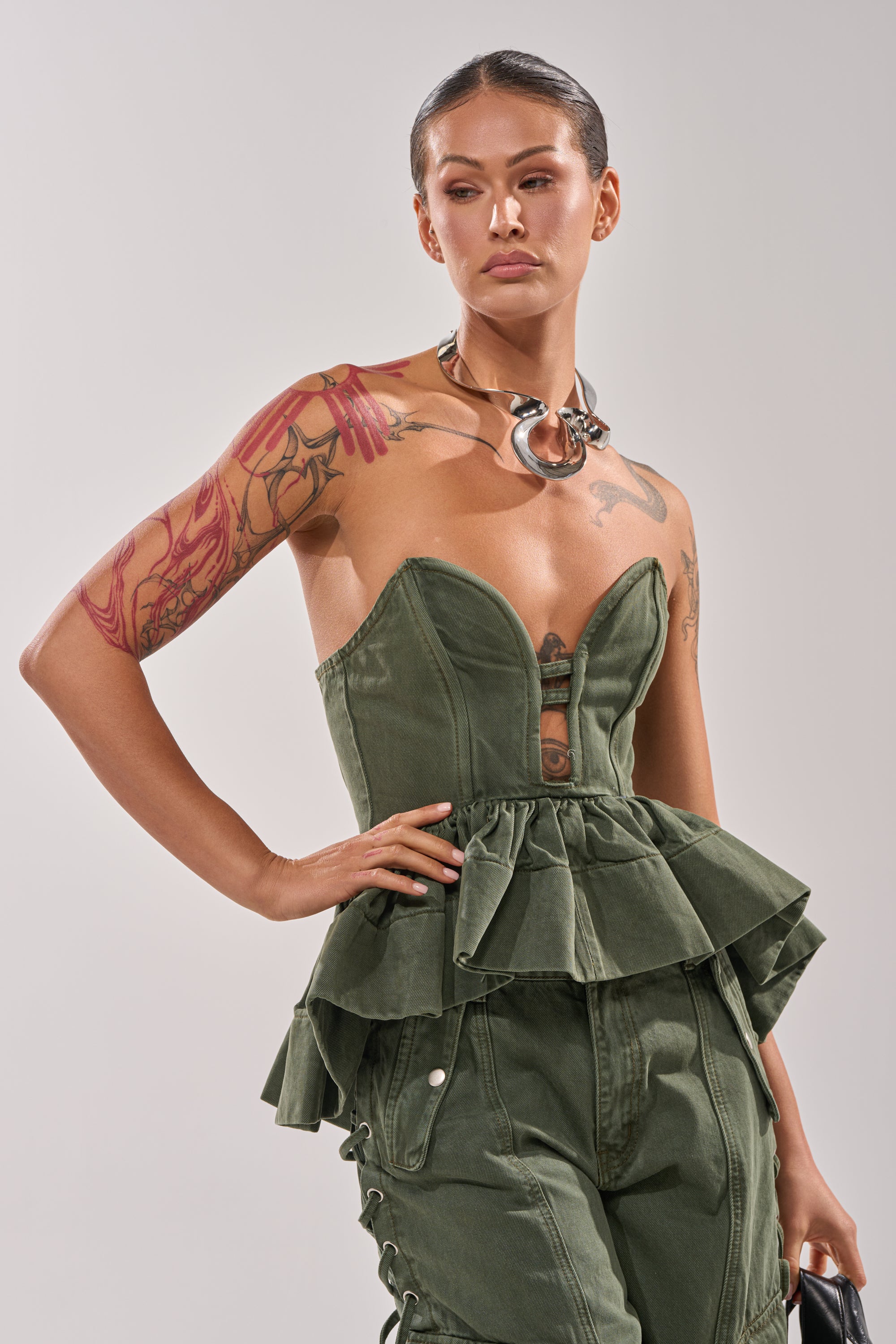 A person with slicked-back hair and tattoos wears the KARA DENIM CORSET, styled with matching pants featuring lace-up details, a chunky metallic choker, and a handbag. 1