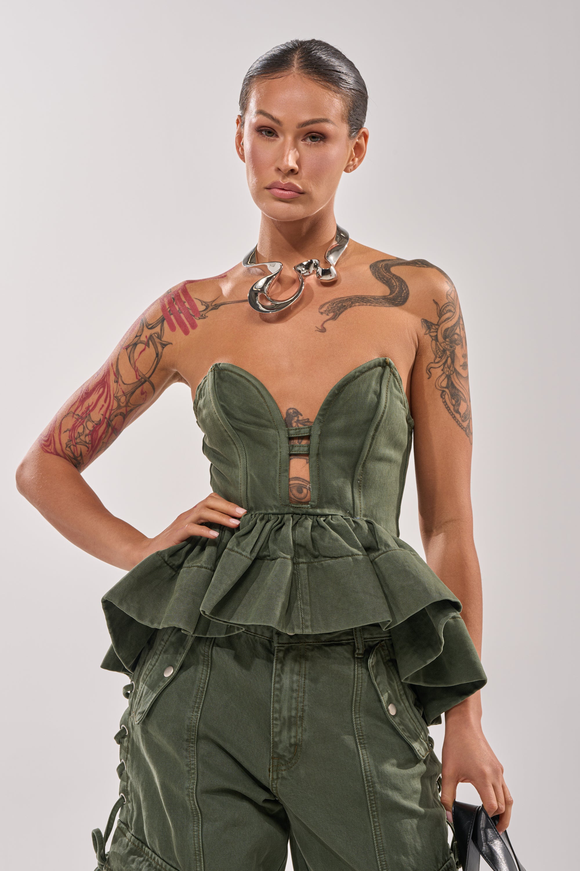 A woman with slicked-back hair wears the KARA DENIM CORSET, matching pants, a chunky metallic necklace, and holds a black bag. She has arm and shoulder tattoos and stands against a plain background. 2