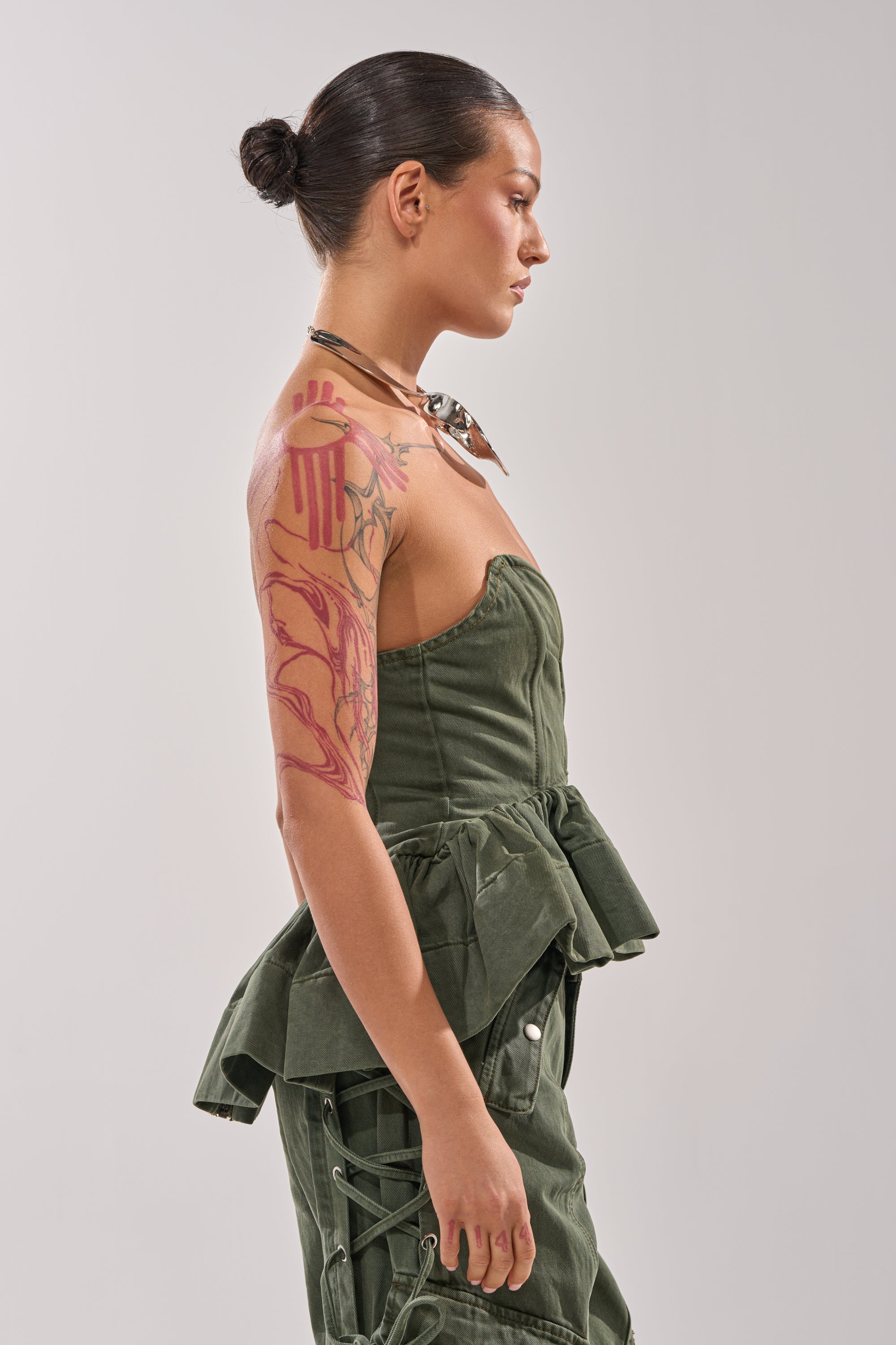 A woman with slicked-back hair in a bun wears the KARA DENIM CORSET and matching pants, accessorized with a choker and showcasing a colorful tattoo sleeve, standing in profile against a plain background. 3