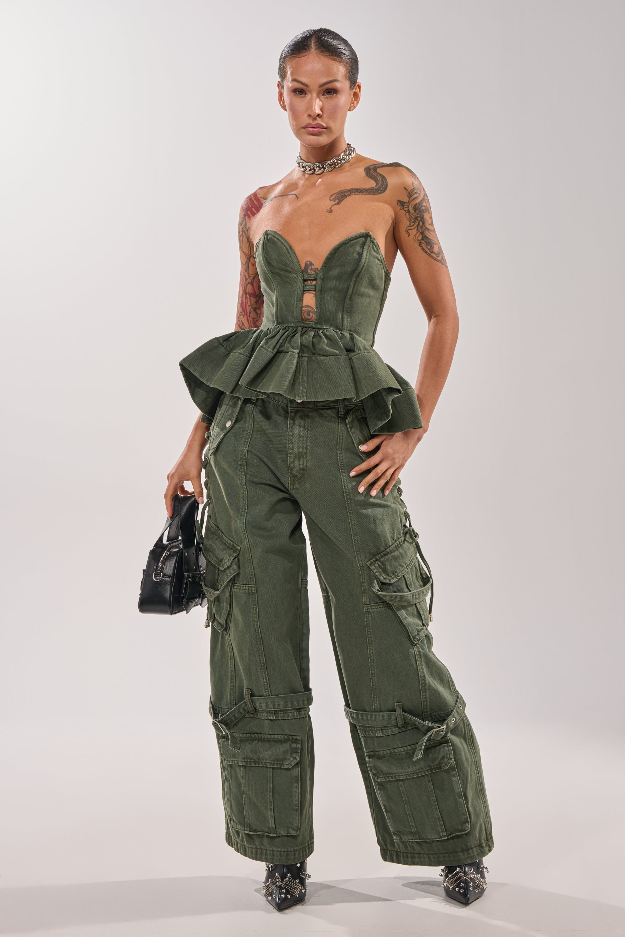 A woman poses confidently in the KARA DENIM CORSET, styled with matching cargo pants, a black handbag, choker necklace, pointed black heels, and showcasing her tattoos. 5
