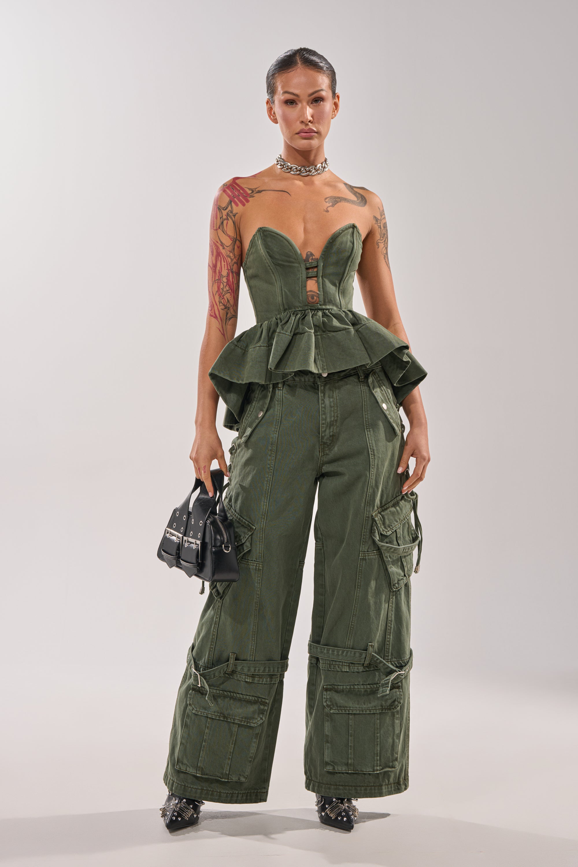 She stands confidently in the KARA DENIM CORSET with a structured sweetheart neckline and matching olive cargo pants, accessorized with a black bag, slicked-back hair, visible tattoos, and black heels with silver spikes. 6