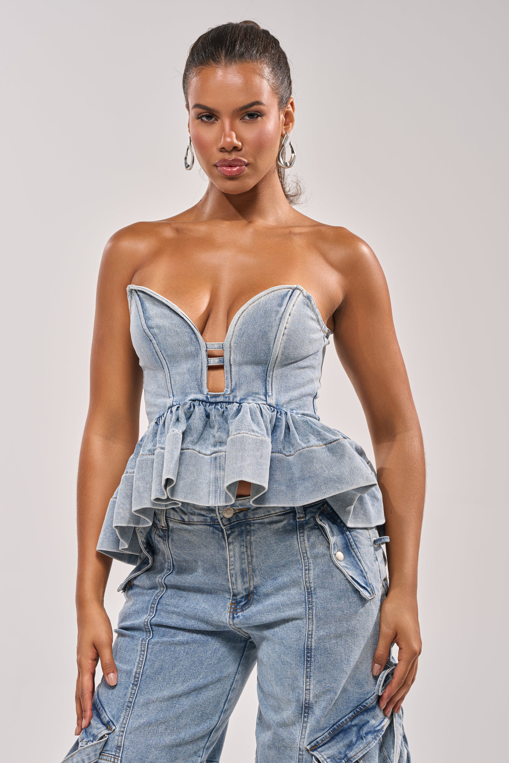 A woman poses confidently in the KARA DENIM BLOUSE, a strapless sweetheart denim peplum top with ruffle details, paired with light blue cargo pants. She accessorizes with hoop earrings and a sleek pulled-back hairstyle for a chic festival look. 1