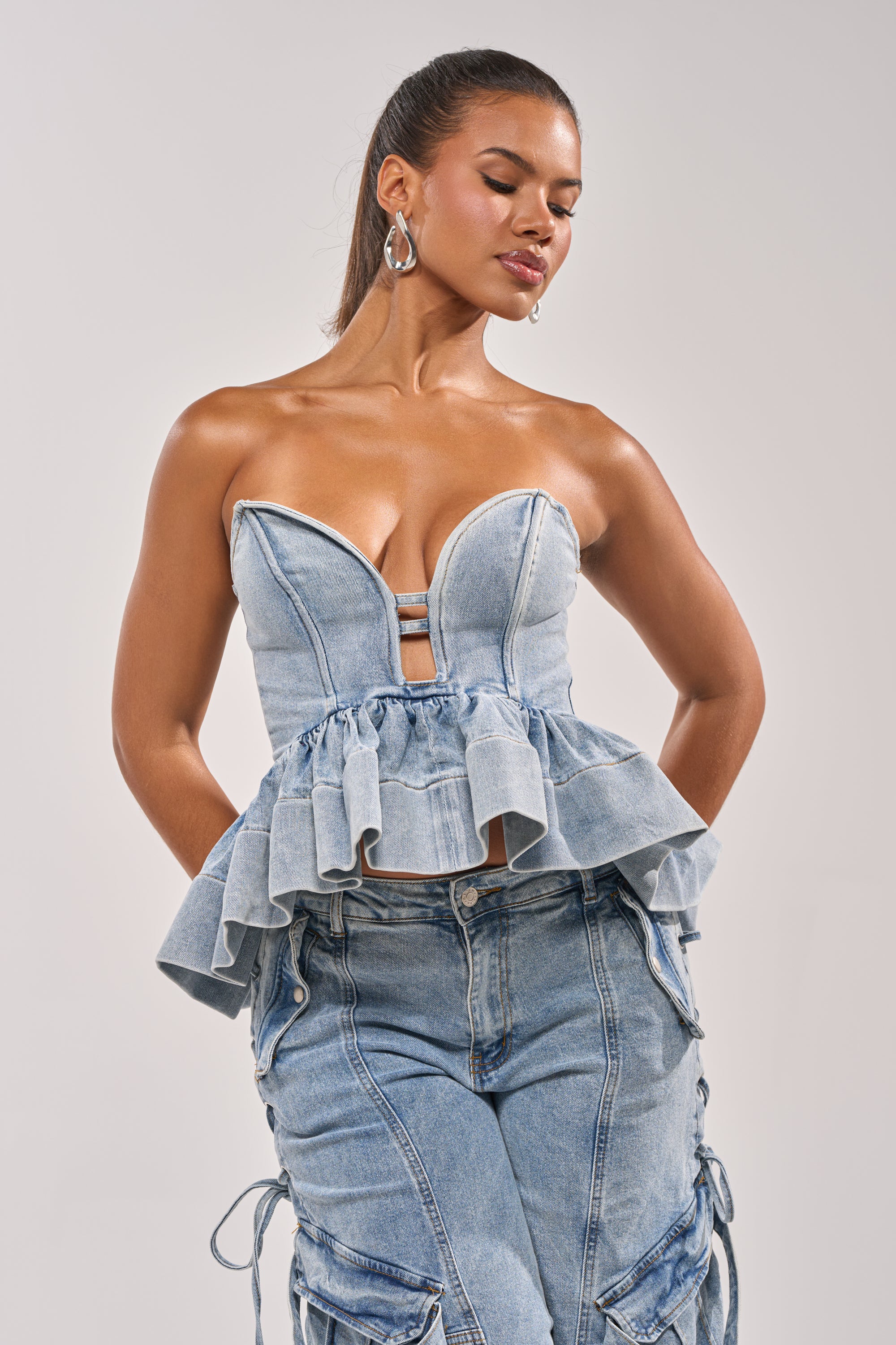 A woman models the KARA DENIM BLOUSE with high-waisted denim cargo pants, hands in pockets and gaze downward—showcasing a stylish, festival-ready look against a plain background. 2