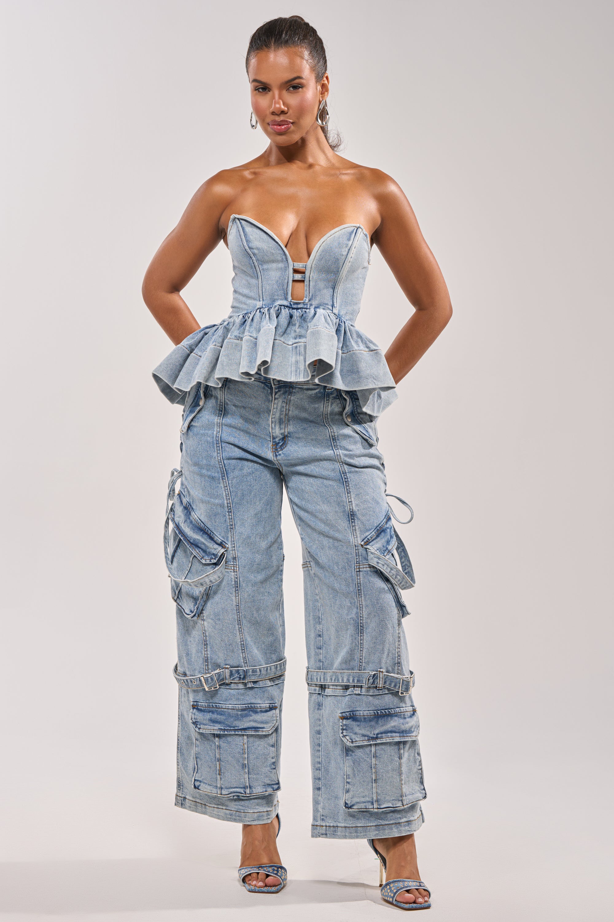 A woman poses confidently in the KARA DENIM BLOUSE—a strapless sweetheart denim peplum top—paired with loose-fit cargo jeans and open-toe heels, crafting a perfect festival look against a plain backdrop. 5
