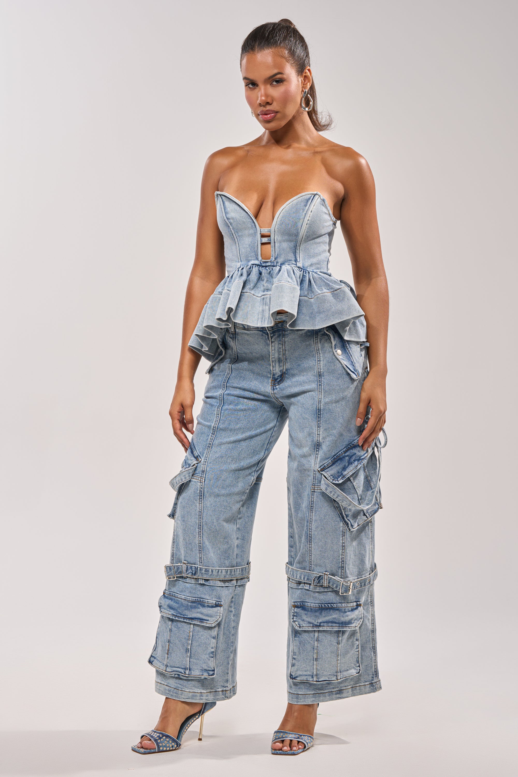 A woman poses against a plain background in the KARA DENIM BLOUSE with a peplum hem and wide-leg cargo denim pants, styled with high-heeled sandals for an effortlessly cool festival look. 6