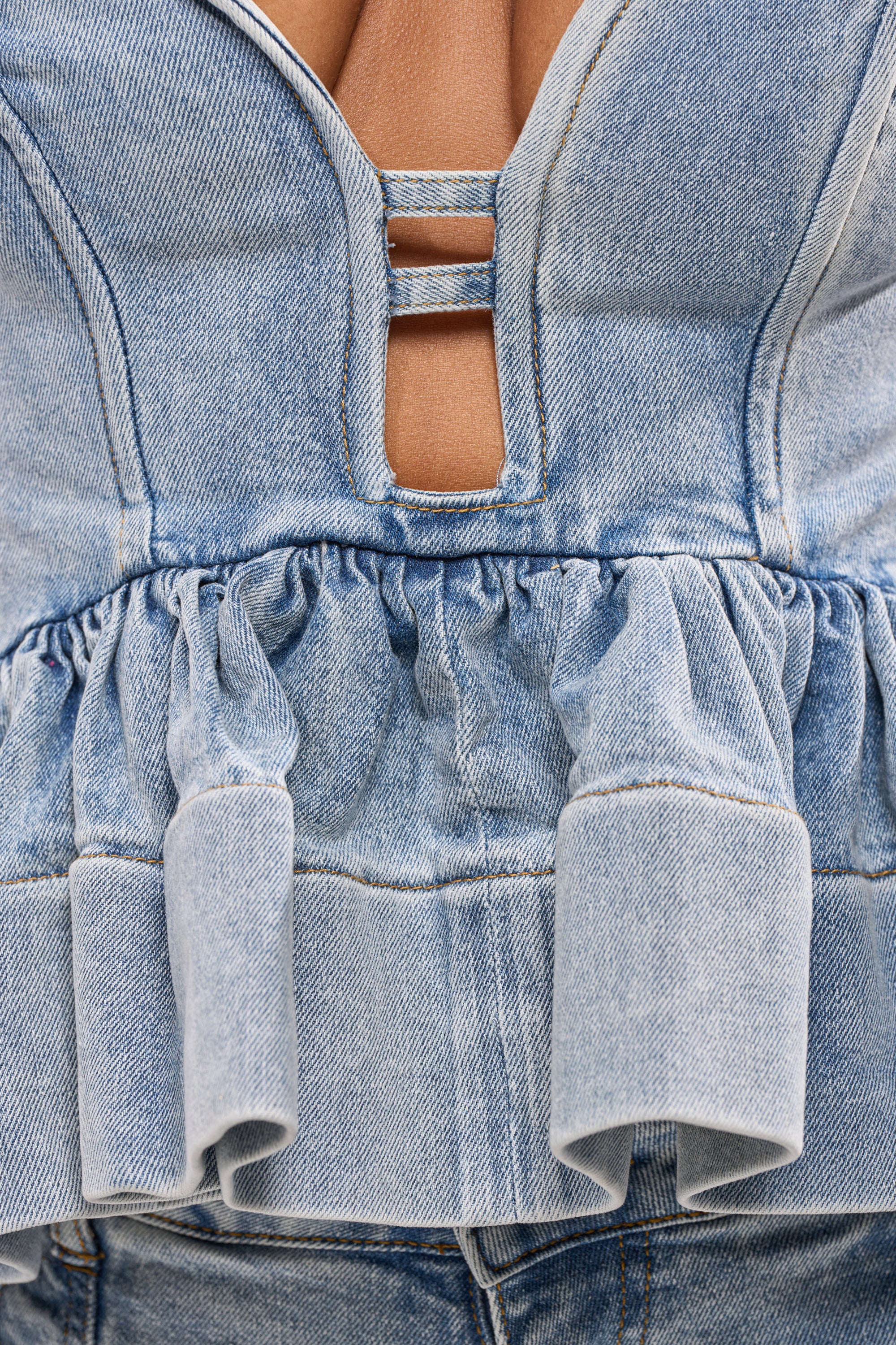 Close-up of the KARA DENIM BLOUSE: a light blue peplum top with structured seams, chest cutout with two horizontal straps, and ruffled hem. Pairs perfectly with matching jeans for the ultimate festival look. 7