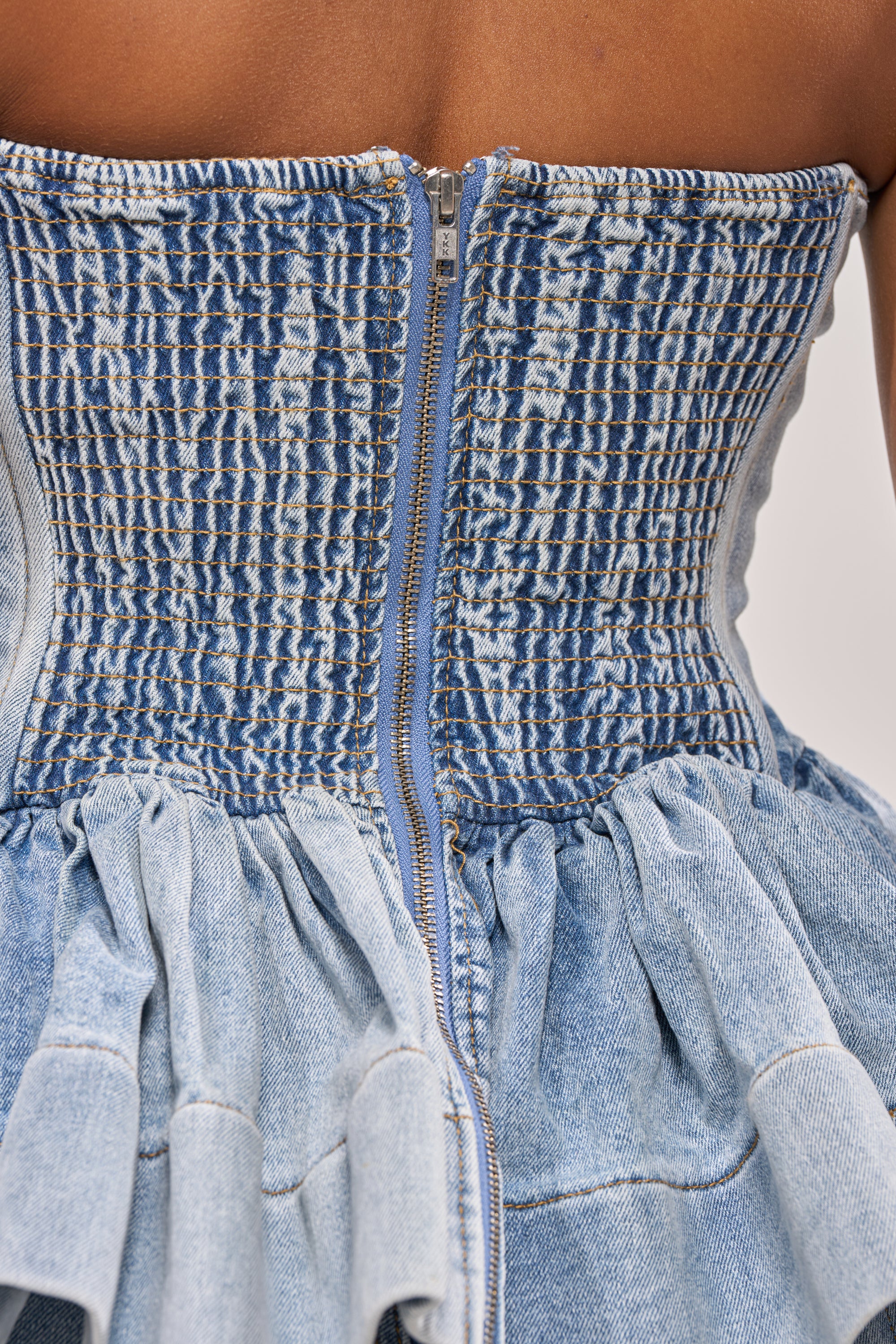 Close-up of the back of the KARA DENIM BLOUSE, a strapless sweetheart top with a visible zipper, smocked back, and gathered ruffled waist—ideal for festivals. Shown on a model with dark skin. 8
