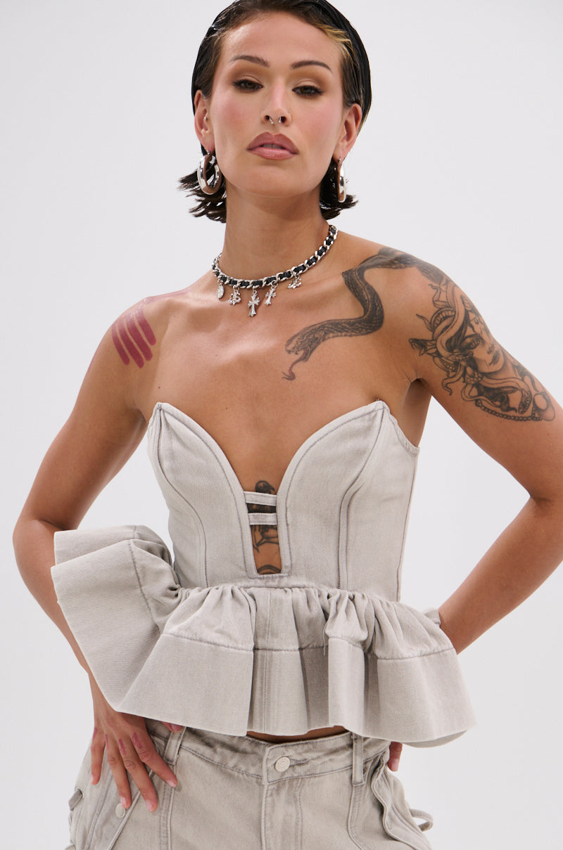 A person with short dark hair and tattoos wears the KARA DENIM BLOUSE, a strapless light gray top with a ruffled peplum hem, paired with matching pants. They accessorize with hoop earrings, a cross necklace, and exude confidence.