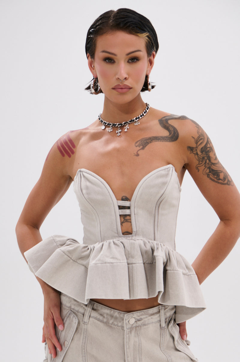 A person with short dark hair wears the KARA DENIM BLOUSE—strapless, structured, with a ruffled peplum hem—paired with matching pants. They show visible tattoos and accessorize with bold earrings and a chunky necklace.