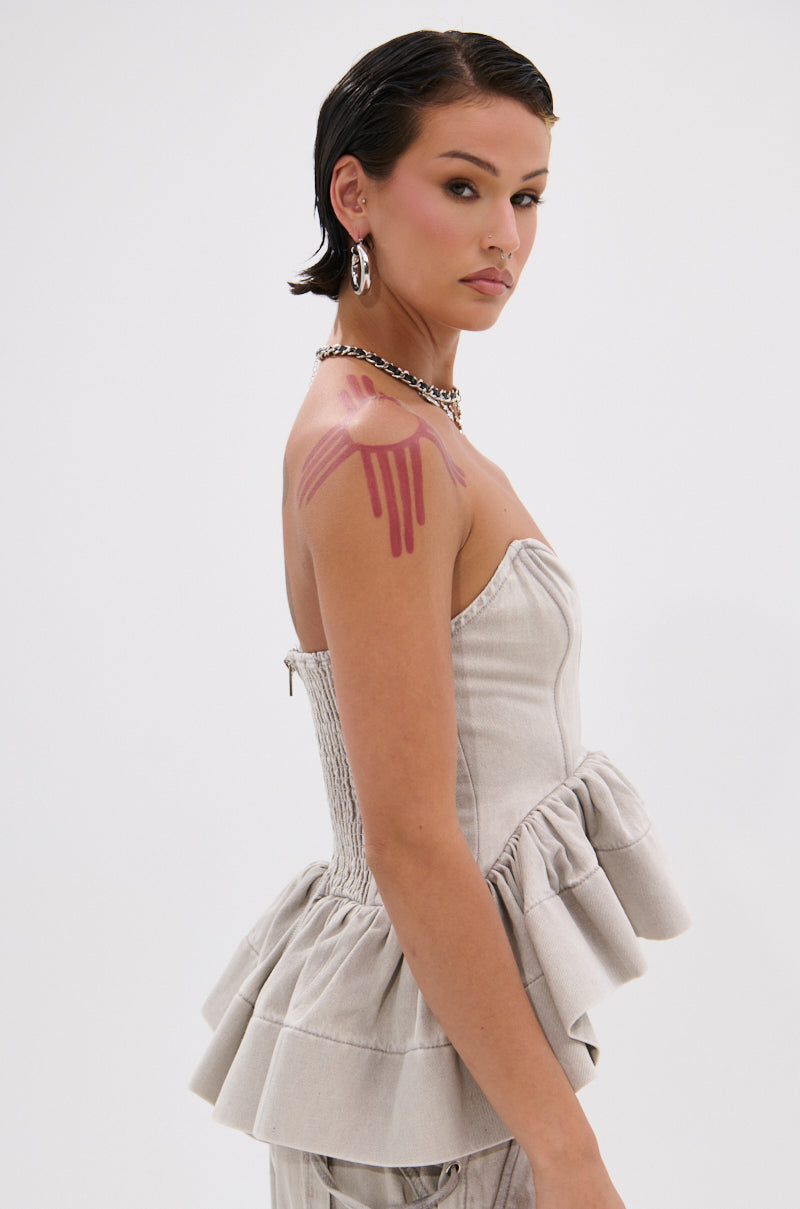 A woman with short dark hair and pink blush faces sideways, wearing the KARA DENIM BLOUSE—a strapless light gray top with a ruffled peplum hem—paired with a chunky chain necklace, hoop earrings, and pink paint dripped on her shoulder.