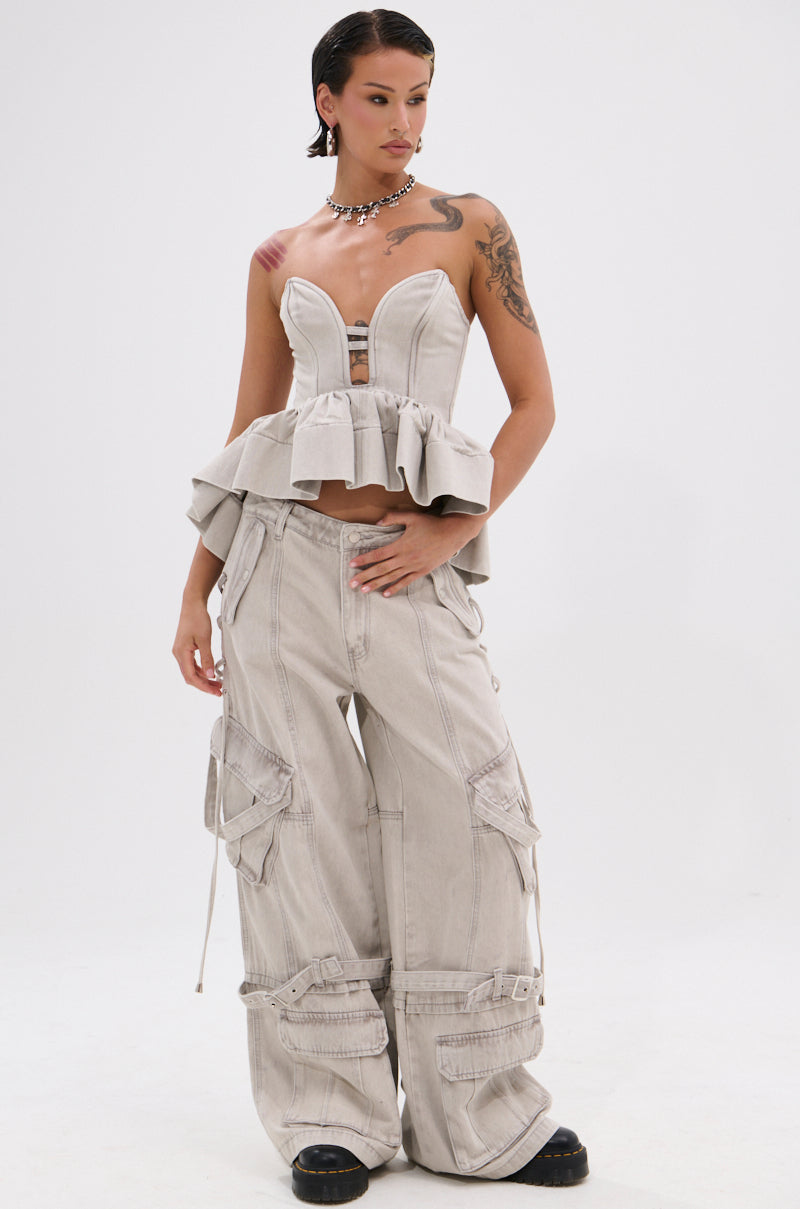 A person wearing the KARA DENIM BLOUSE, a strapless peplum top with a sweetheart neckline, and wide-leg light gray cargo pants stands against a plain white background. They have short dark hair, visible tattoos, and wear black chunky shoes.