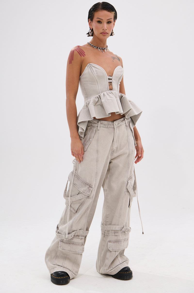 A person stands confidently in the KARA DENIM BLOUSE, a light grey, strapless peplum corset top with a sweetheart neckline, paired with matching wide-leg cargo pants. They have short, slicked-back hair and wear chunky black shoes.