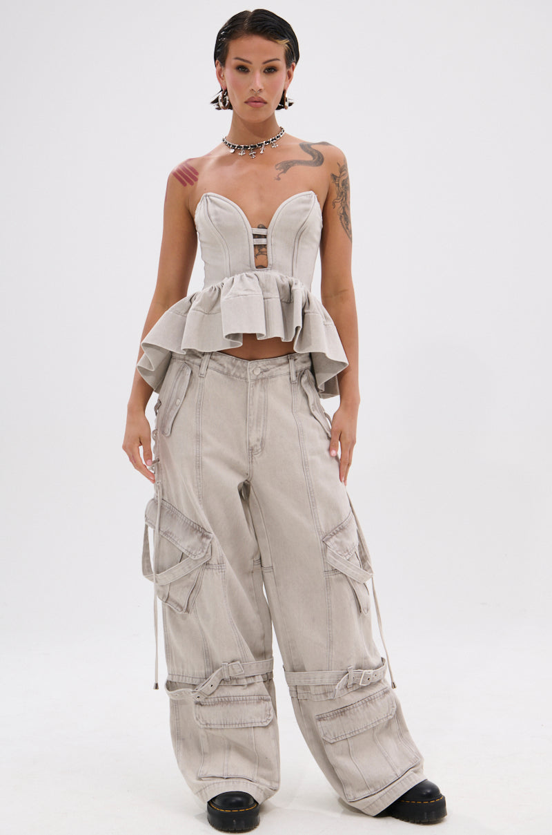 A woman with short dark hair wears the KARA DENIM BLOUSE, a structured strapless top with a sweetheart neckline and ruffled peplum hem, paired with loose cargo pants featuring multiple pockets and zippers against a plain white background.