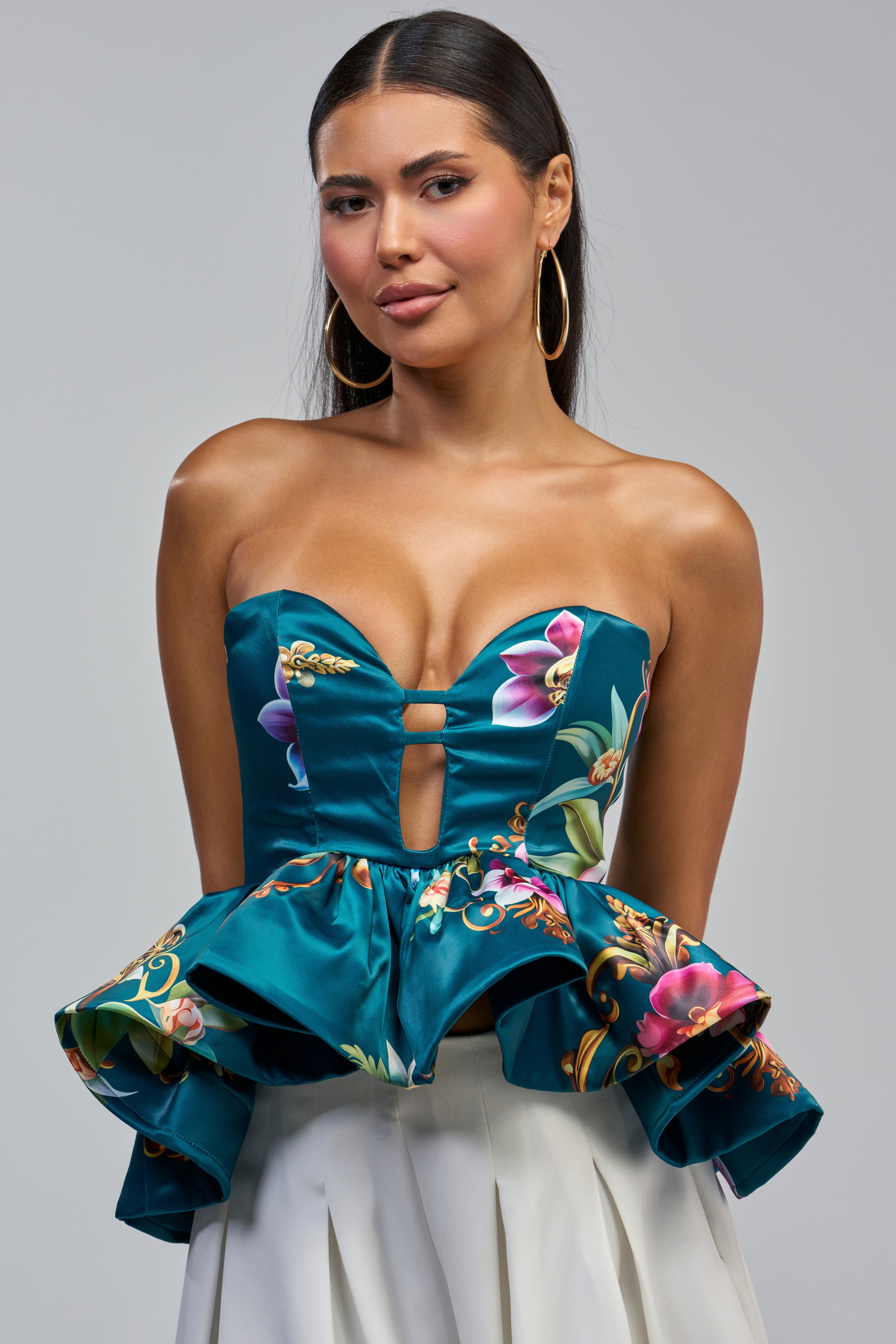 A woman poses against a light gray background wearing the ISLAND TIME FLORAL CORSET, featuring a teal floral print and a strapless sweetheart neckline, paired with gold hoop earrings and a white pleated skirt.