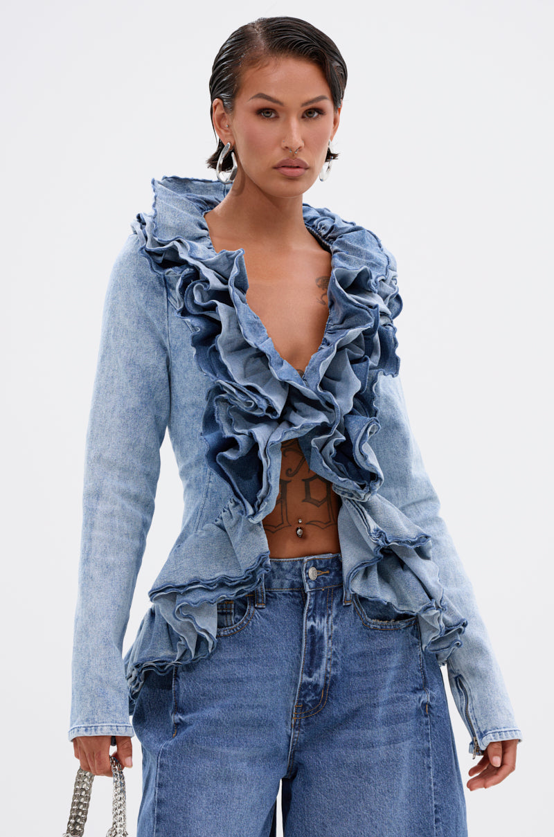 Wearing the WILD WEST RUFFLE DETAIL BLOUSE in light blue denim with dramatic ruffles, paired with high-waisted jeans, hoop earrings, and a chain handbag, a confident model poses against a plain white background.