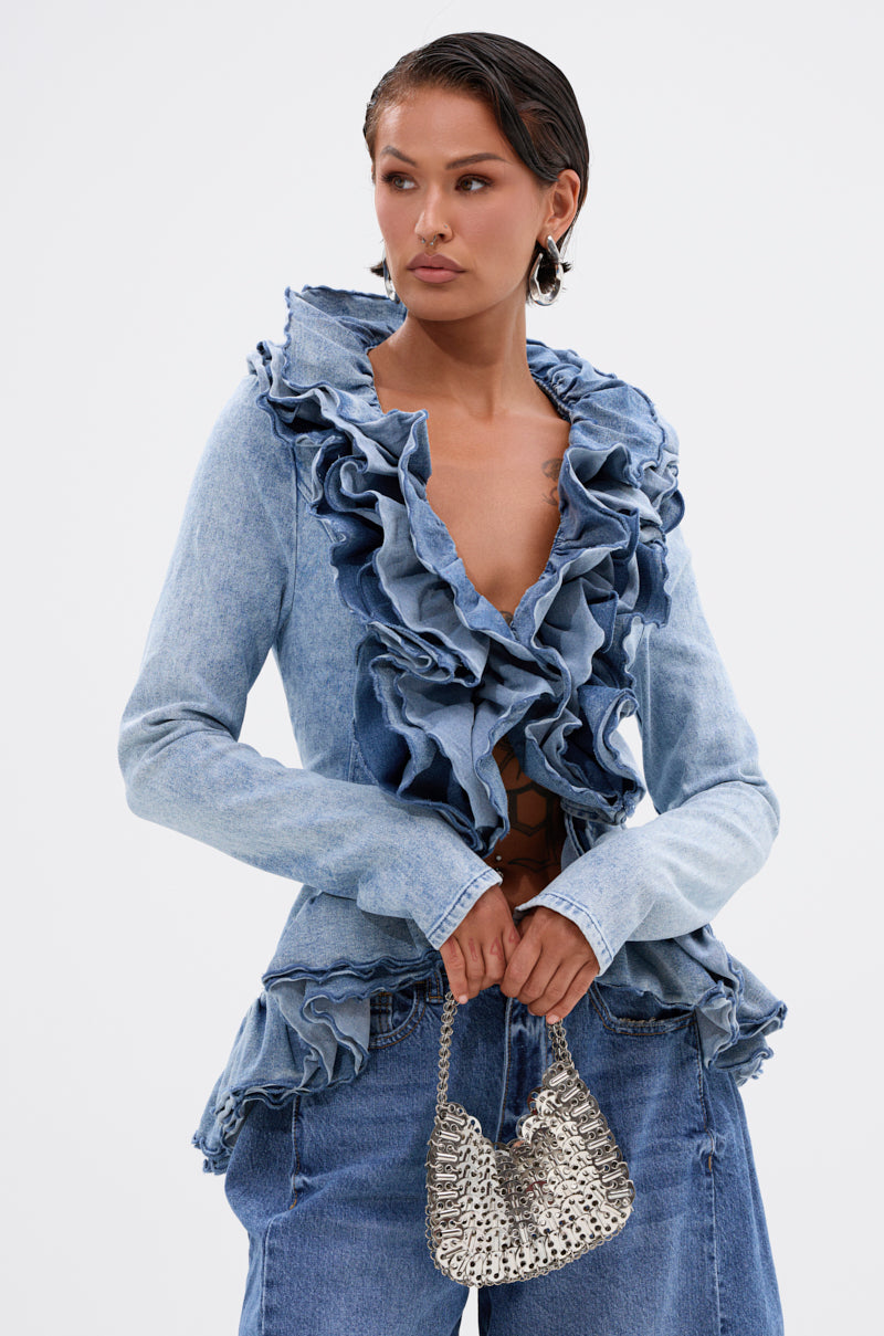 A person wears the WILD WEST RUFFLE DETAIL BLOUSE in light blue denim with dramatic ruffled edges, paired with matching jeans and a small silver textured handbag against a plain white background.