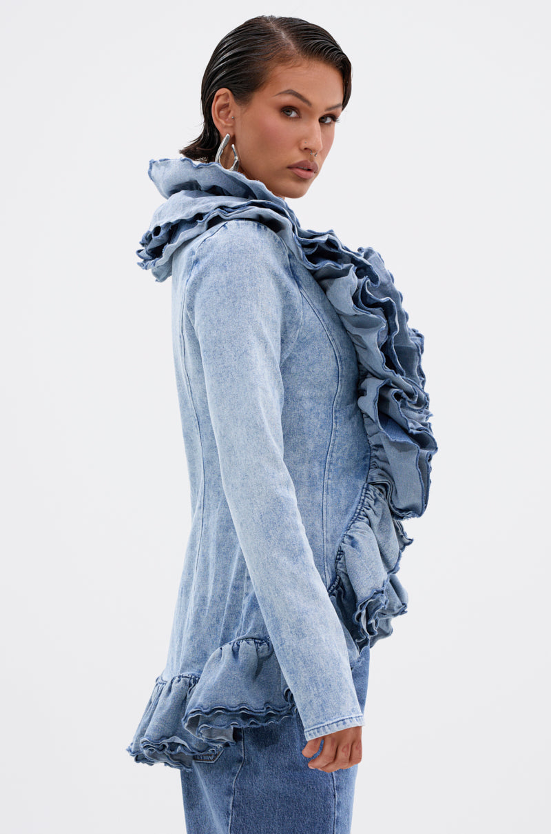 A woman wears the WILD WEST RUFFLE DETAIL BLOUSE in light blue denim, featuring dramatic ruffles on the collar, front, and hem, paired with matching denim pants, posing against a plain white background.