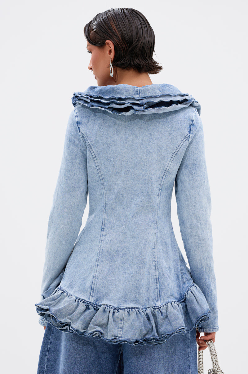 A woman with short, dark wet-look hair wears the WILD WEST RUFFLE DETAIL BLOUSE in light blue denim, featuring fitted ruffles at the collar and hem, paired with matching wide-leg jeans. She faces away to showcase the back of the outfit.