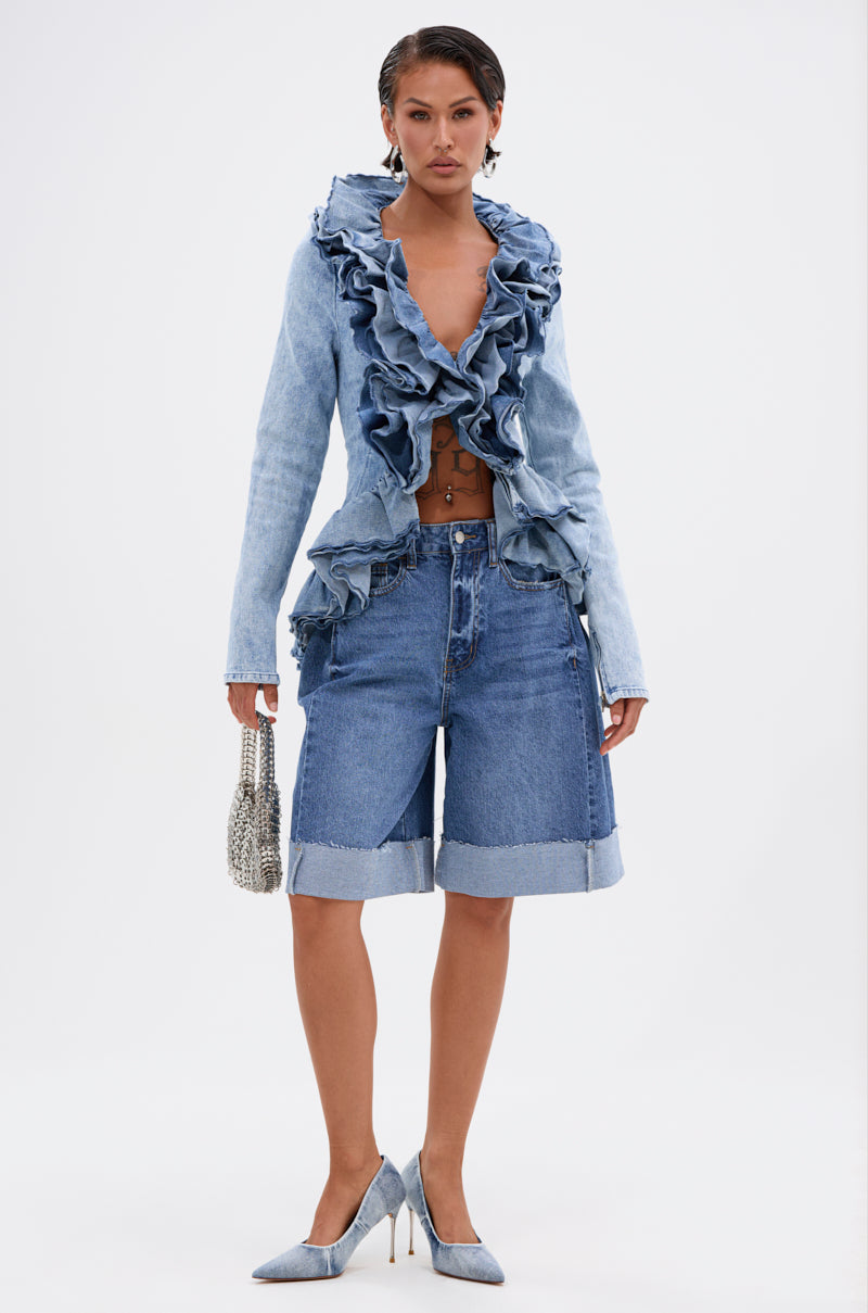 A woman poses confidently against a plain white background, wearing the WILD WEST RUFFLE DETAIL BLOUSE in light blue denim, styled with rolled-up knee-length denim shorts, pointed blue heels, and a small silver handbag.